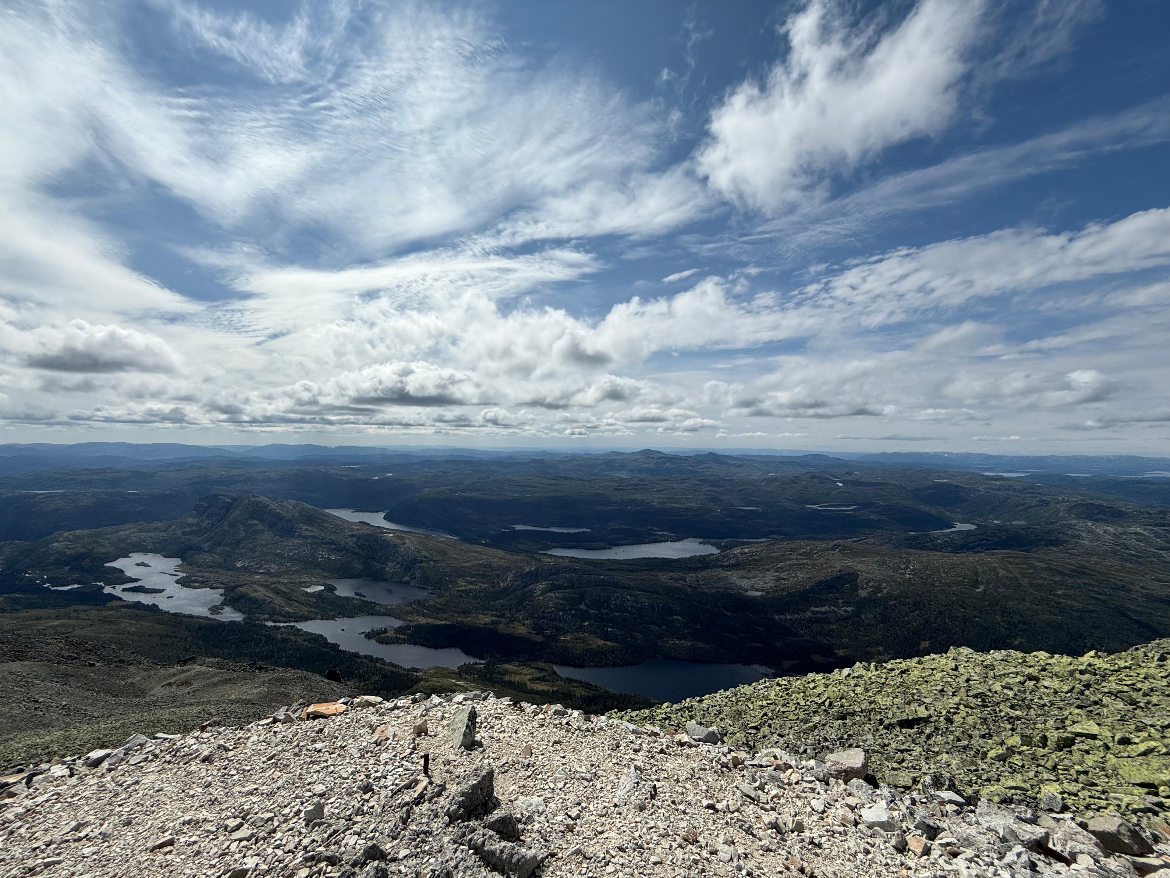 Vidt utsyn fra steinete fjelltopp over skog, vann og fjell under skyer.