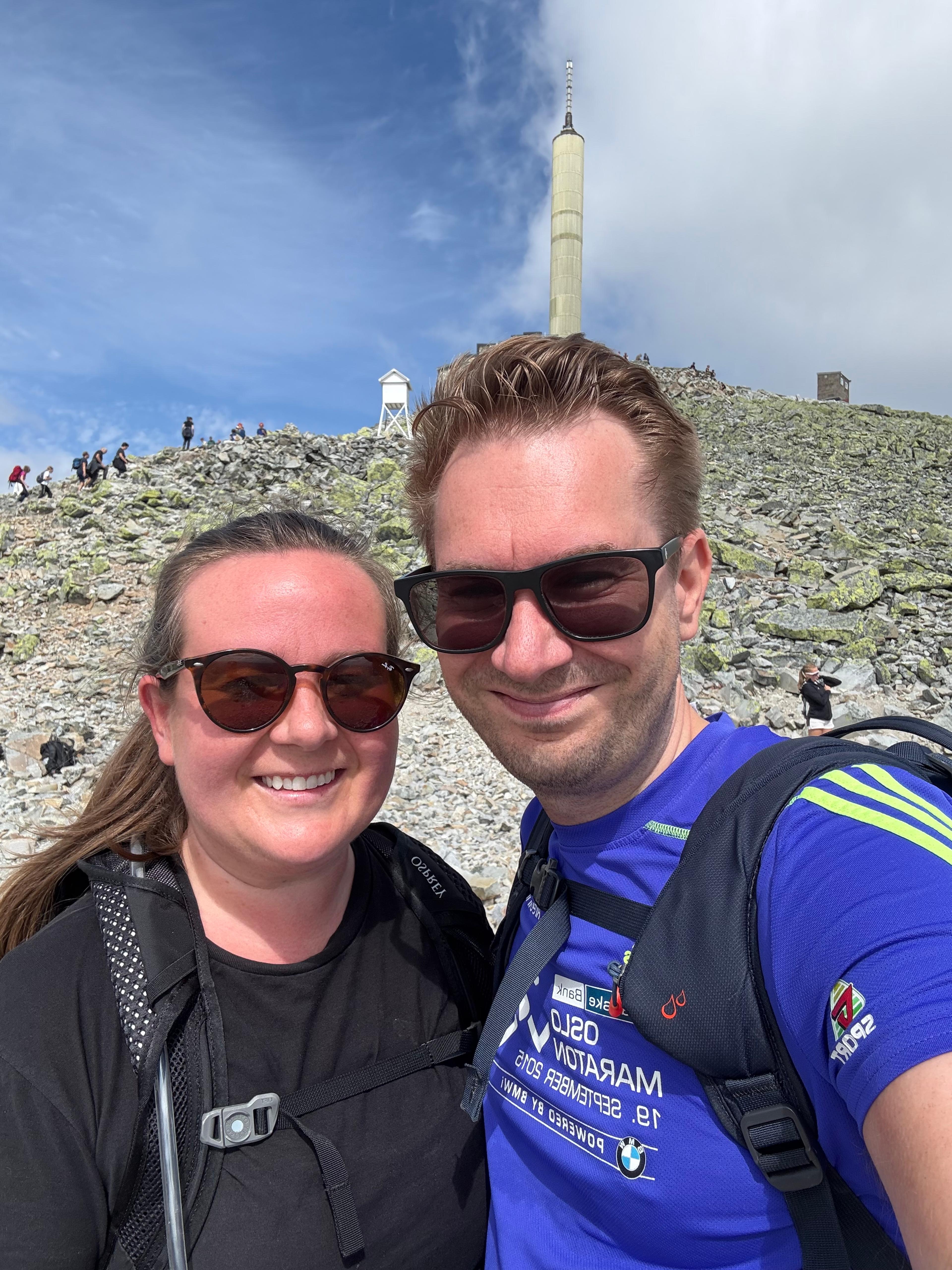 Smilende par med solbriller tar selfie på steinete topp, med tårnet og mange turgåere bak seg.