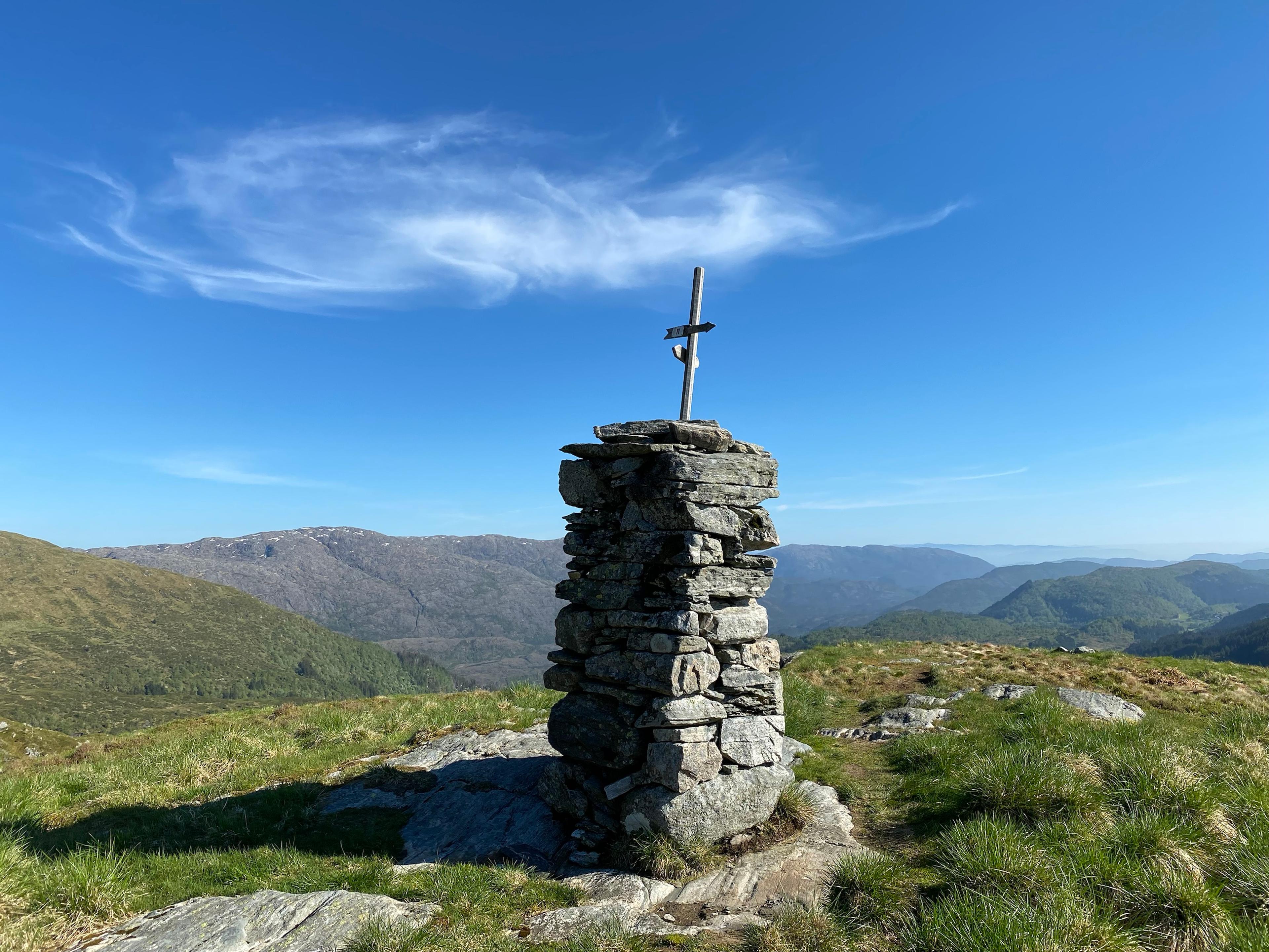 Høy steinvarde med en trestolpe og veiskilt på toppen, på et gresskledd fjellplatå med blankskurt berg, og fjell og daler i bakgrunnen.