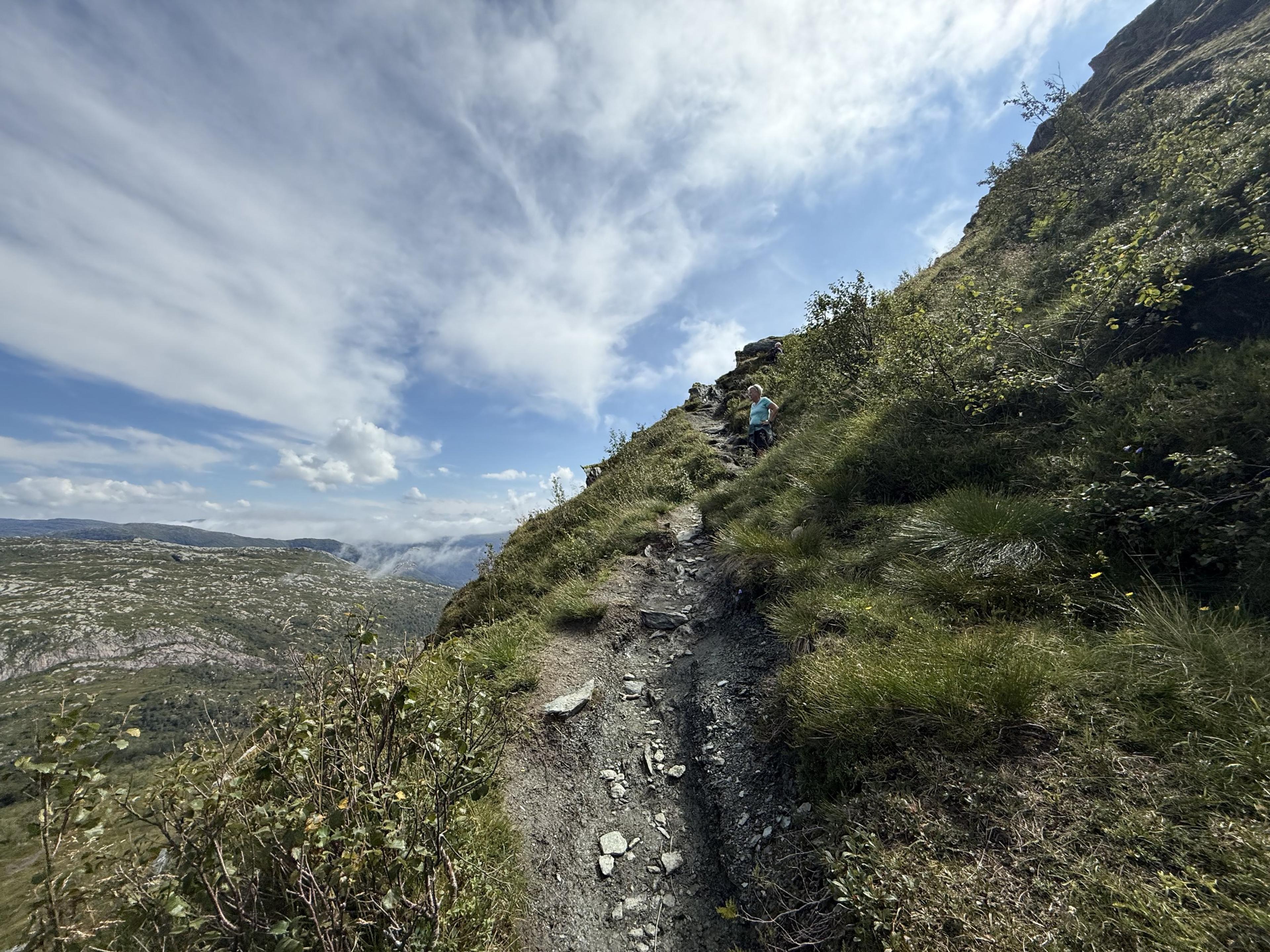 Smal, steinete sti opp en bratt gresskledd fjellside under en blå himmel med slørskyer.