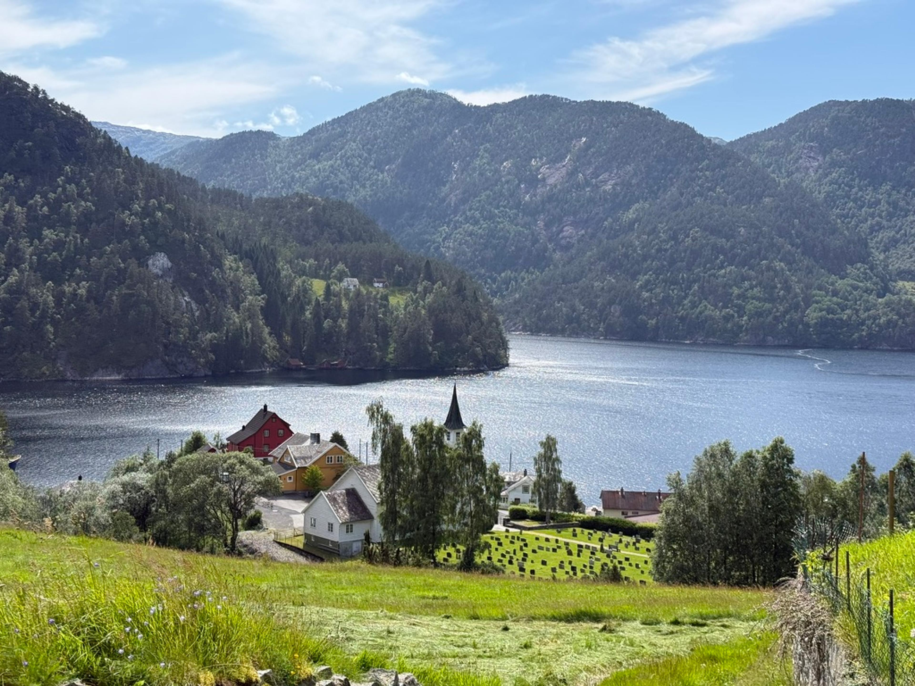 Hvit kirke med spir og en kirkegård ved fjorden, med noen hus rundt og skogkledde fjell på den andre siden.