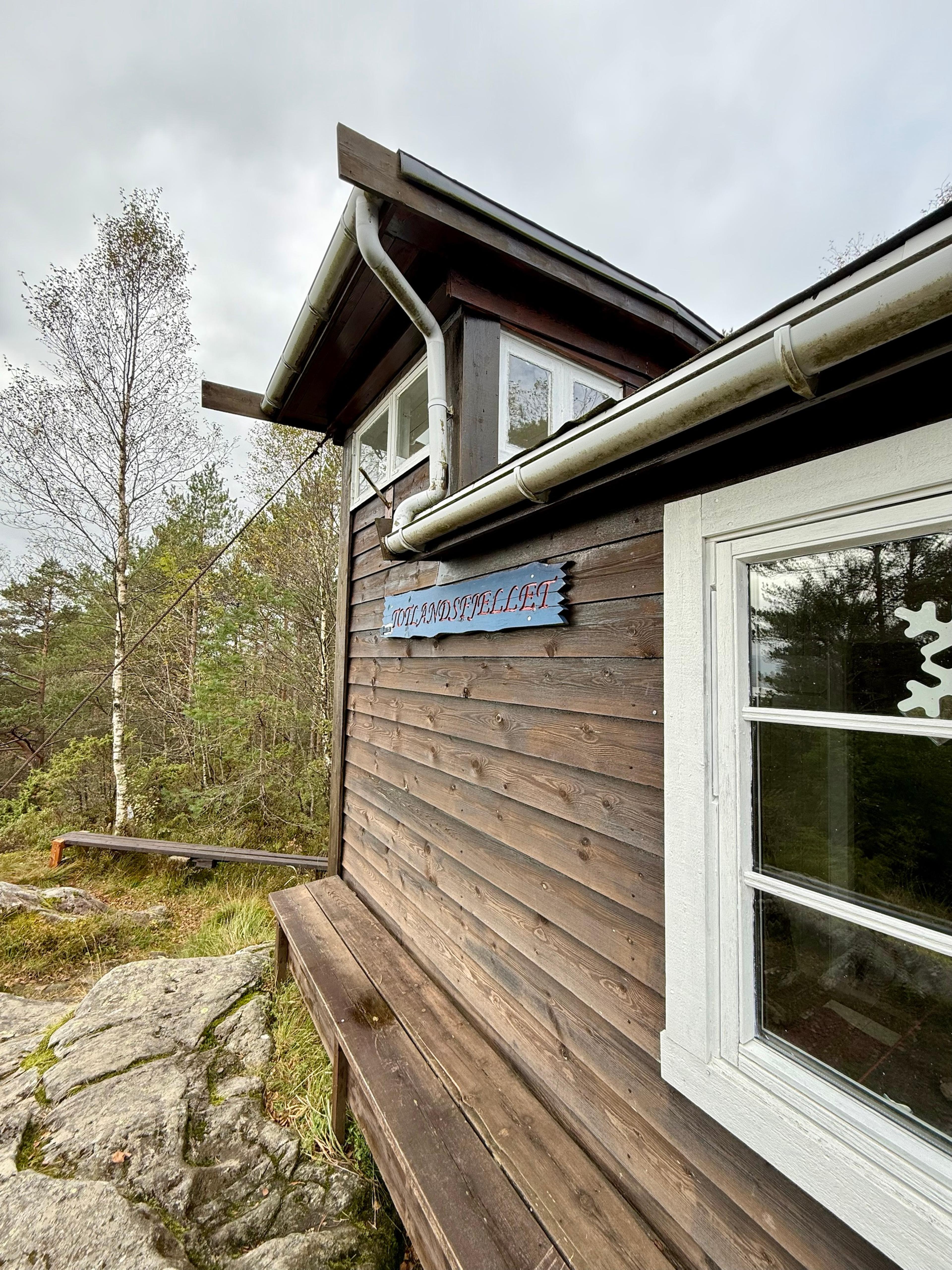 Brun trehytte med hvite vinduer og et blått skilt med teksten «Totlandsfjellet», med en benk langs veggen, svaberg foran og bjørk og furu bak.