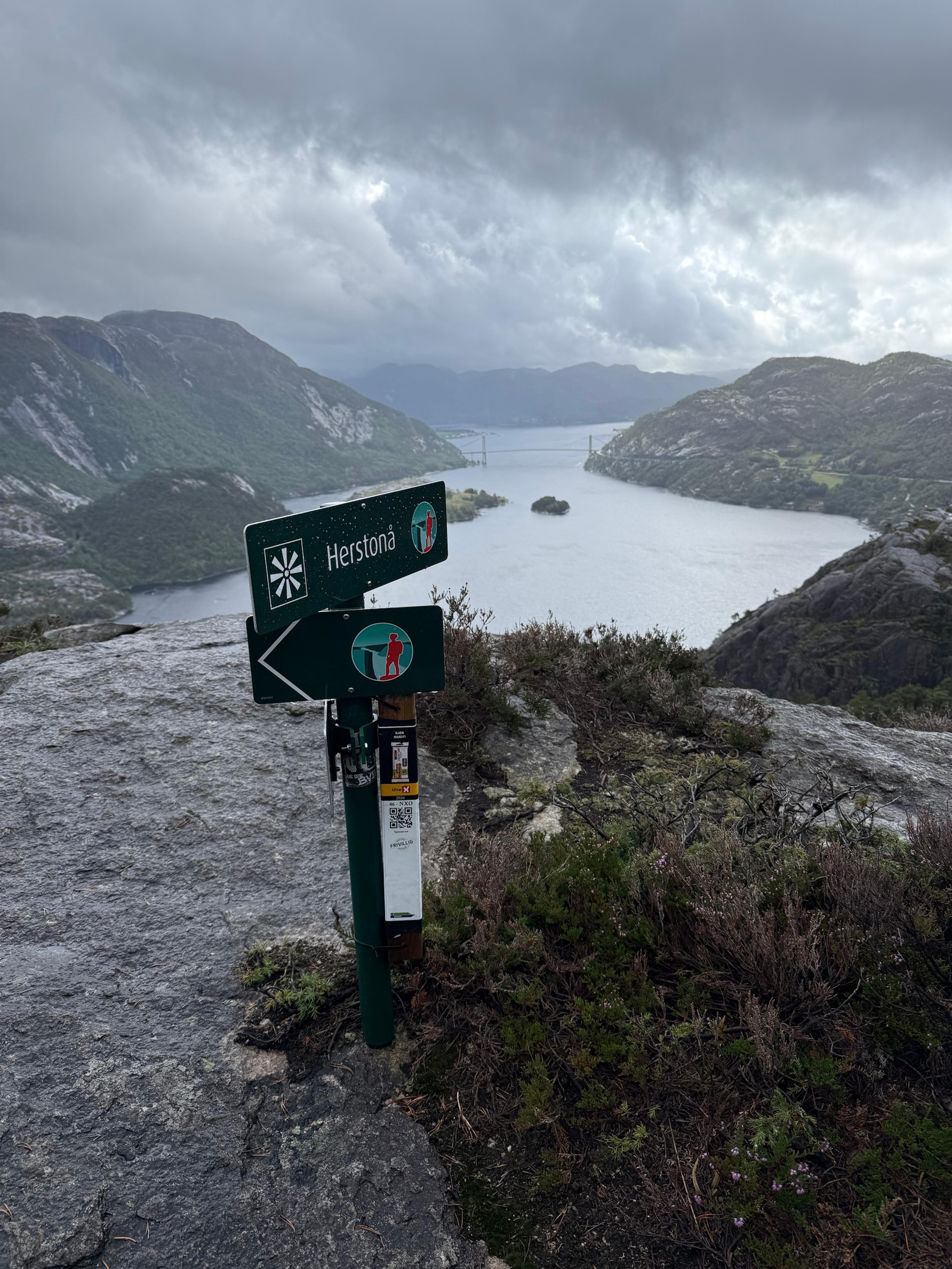 Skilt merket «Herstonå» på toppen, med utsikt ned på Lysefjorden og Lysefjordbrua under tunge skyer.