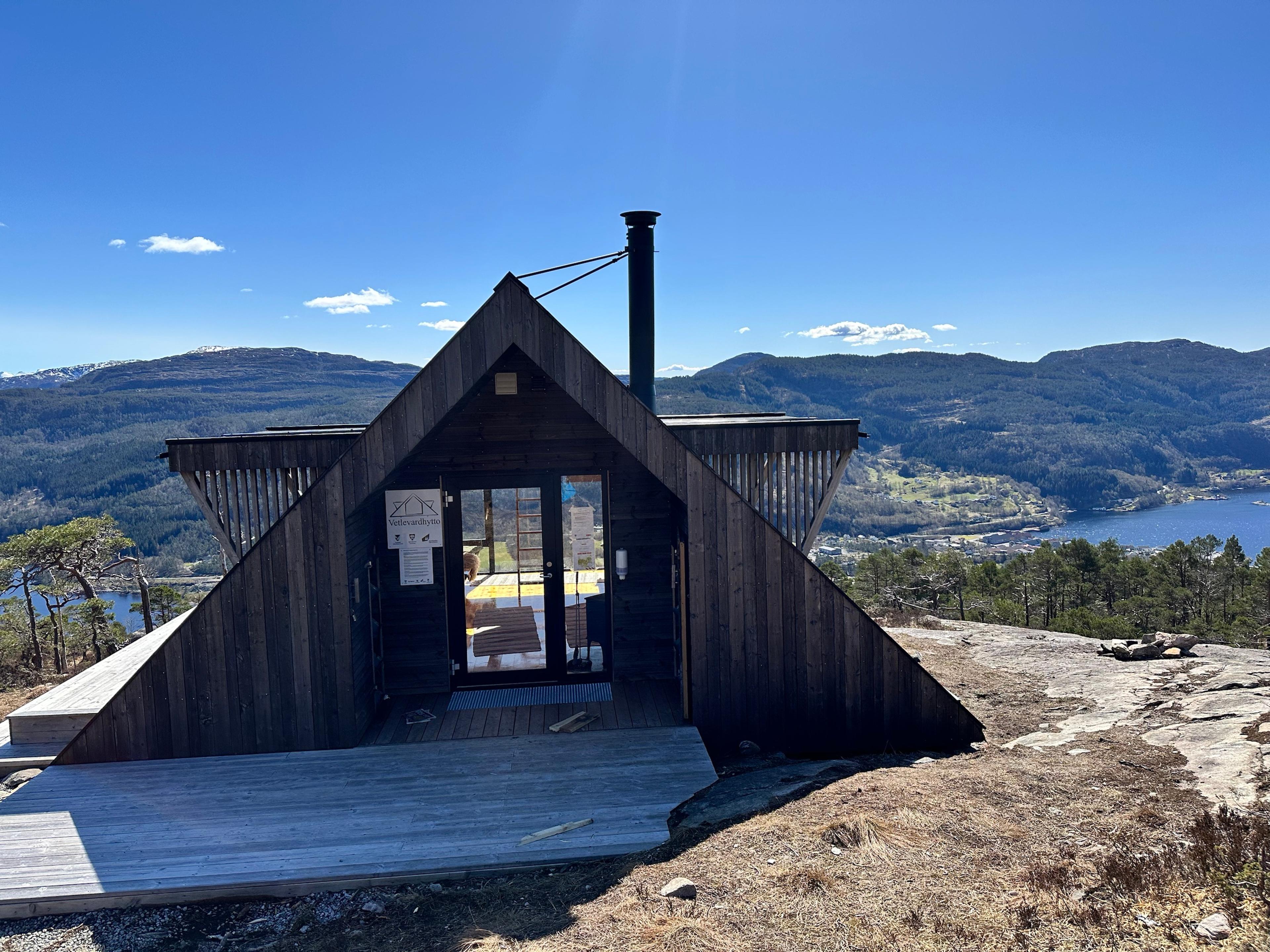 Mørk trehytte med skiltet «Vetlevardhytto» over døren, med fjorden og bygden Eikelandsosen nedenfor.