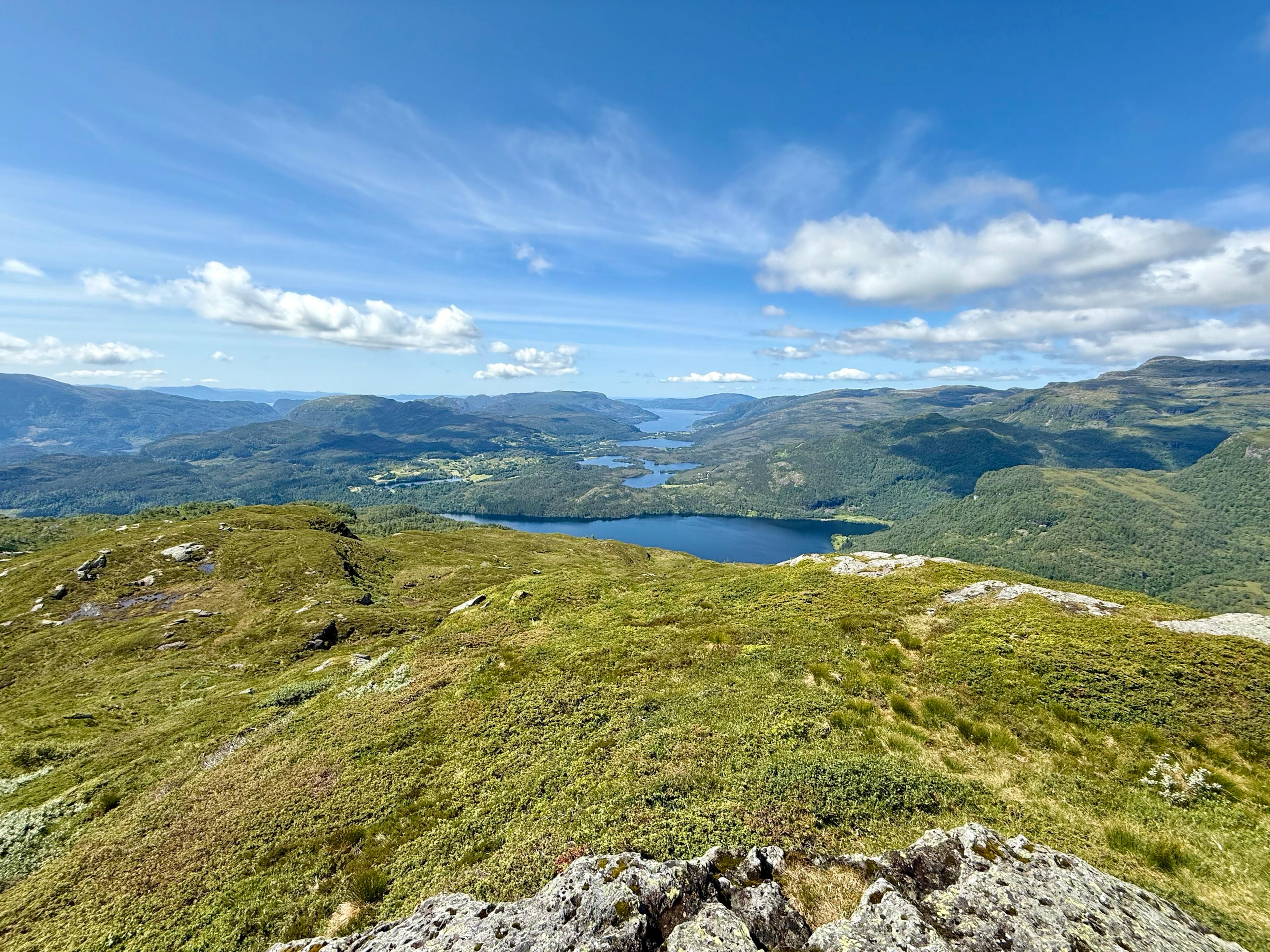 Utsikt fra en gressdekt topp ned på et stort, mørkeblått vann, med flere mindre vann og jorder lenger ute i dalen og en fjord mot horisonten.