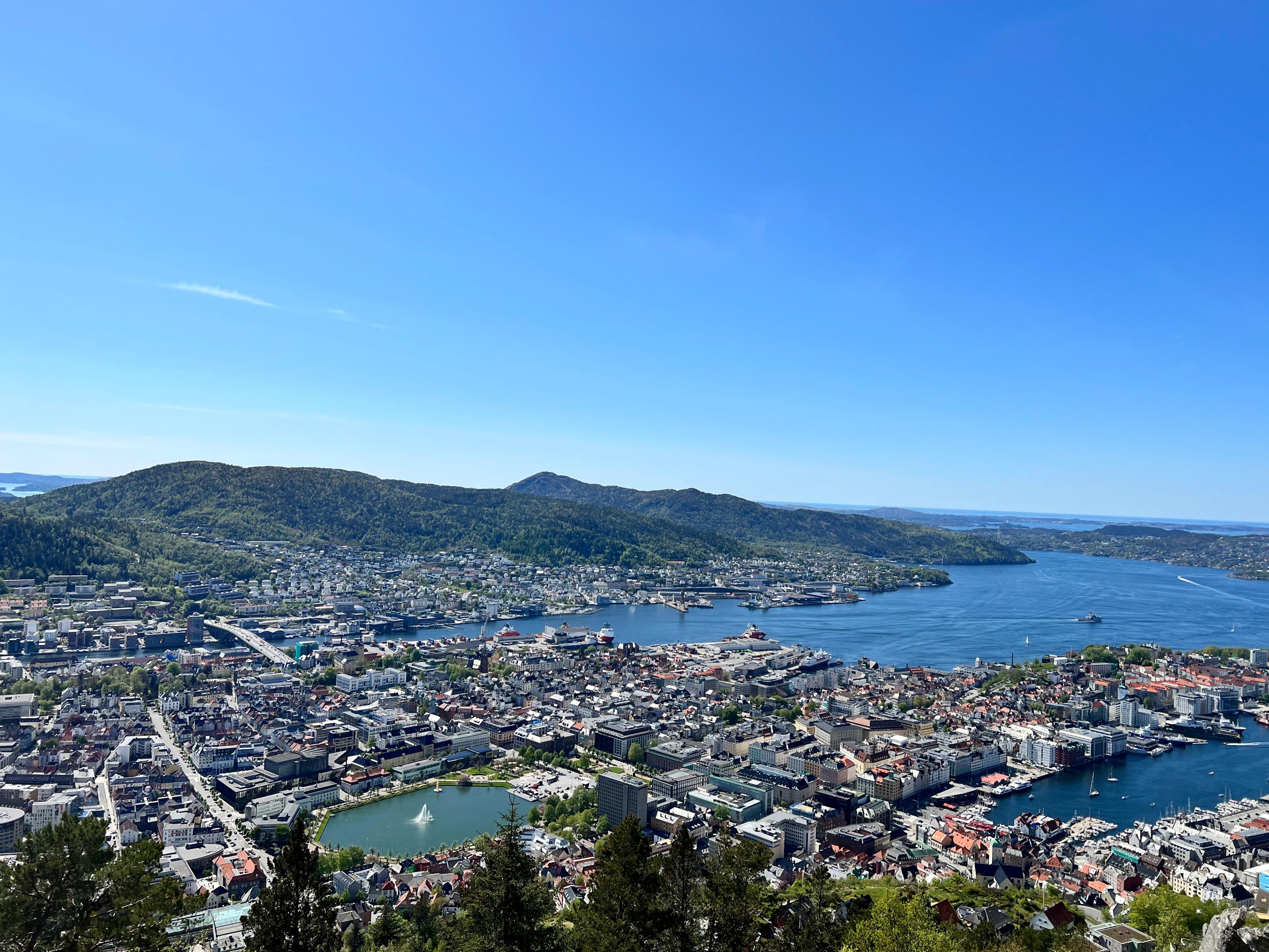 Utsikt fra Fløyen over Bergen sentrum på en skyfri dag. Lille Lungegårdsvann med fontenen ligger midt i bildet, havnen med skip ved kai til høyre, og Byfjorden går ut mot øyene i horisonten.