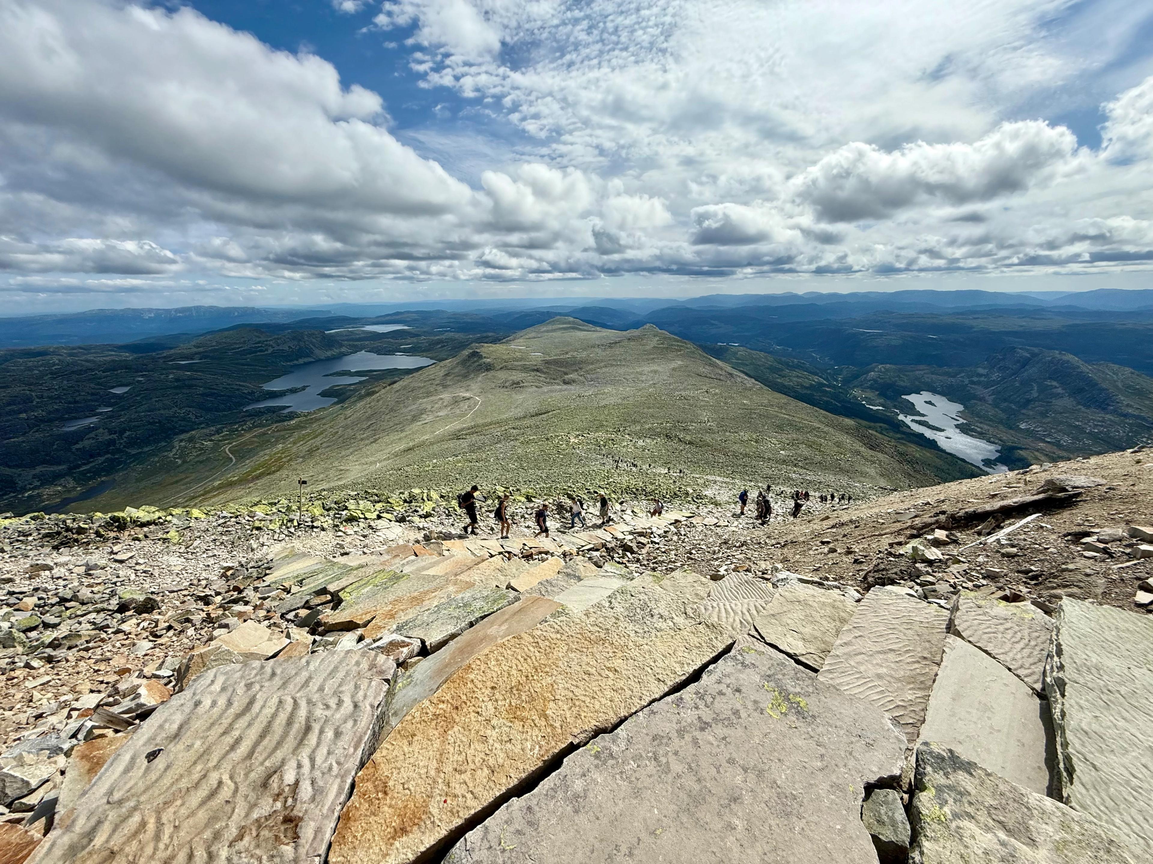 Utsikt fra steinete topp ned en lang fjellrygg med turgåere, med vann og skog på begge sider.