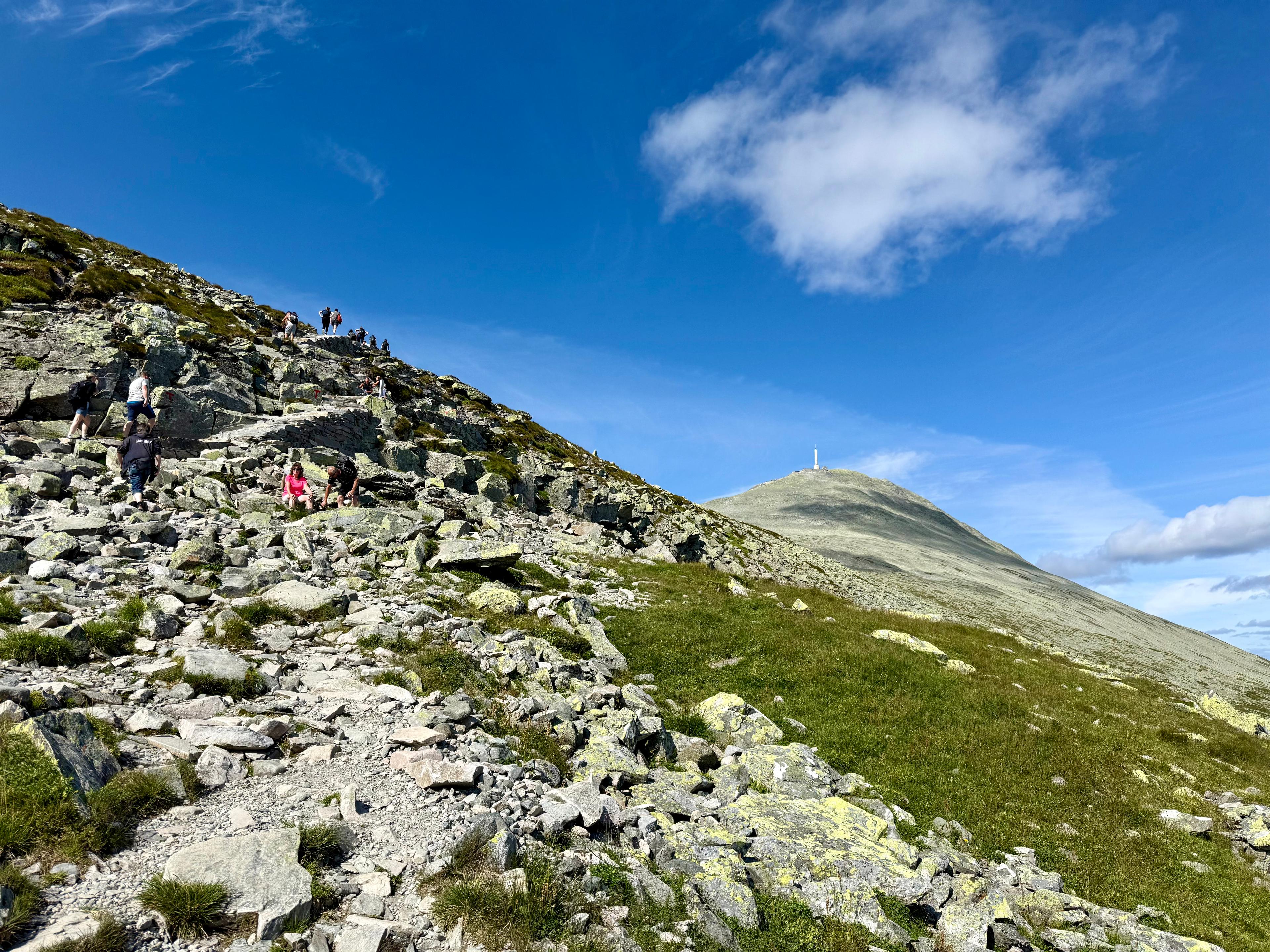 Steinete sti oppover en fjellside med mange turgåere, og Gaustatoppen med tårnet til høyre.