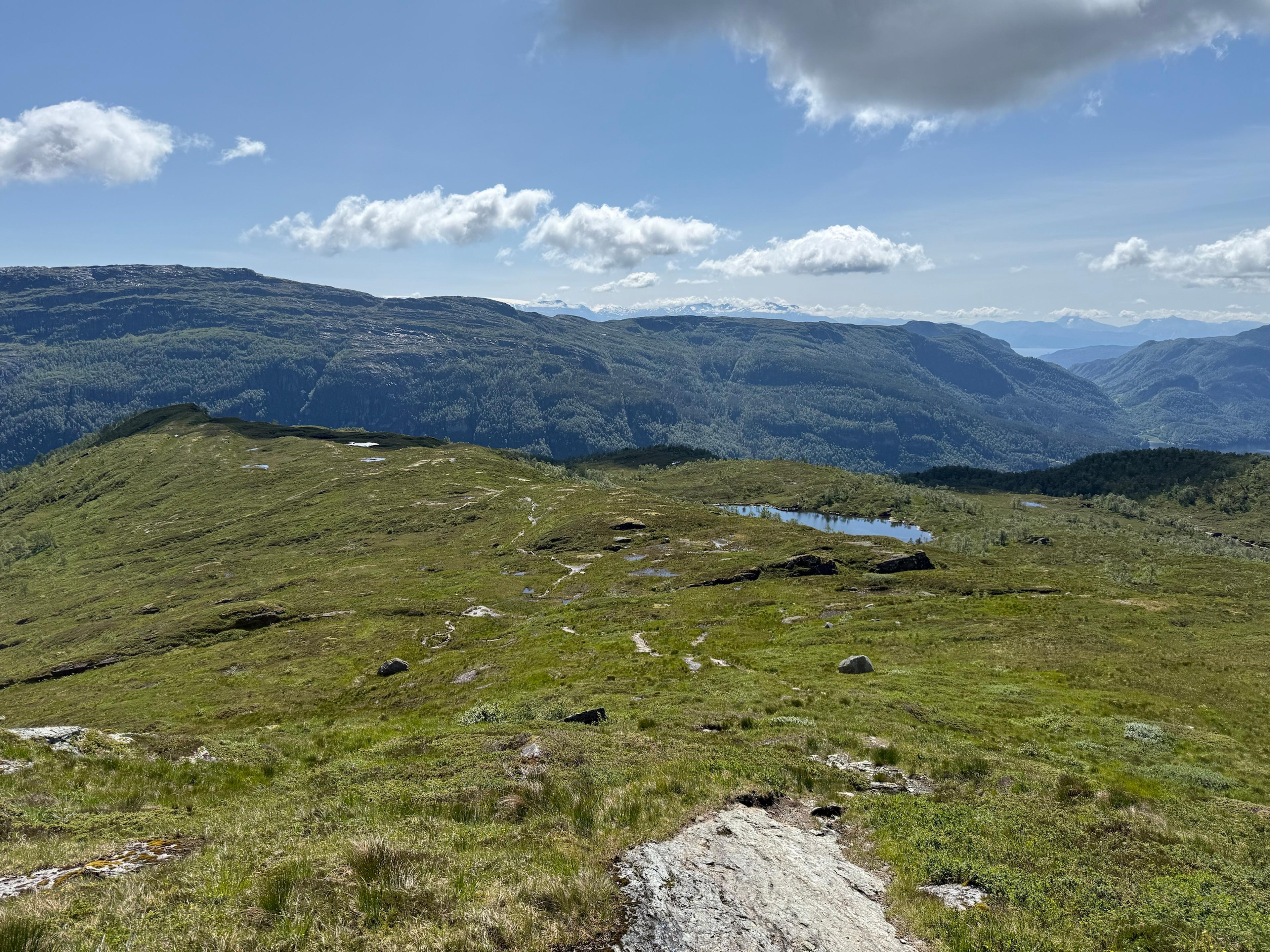 Slakt, gressdekt fjellterreng med svaberg, steiner og en sti som snor seg ned mot et lite tjern. Bak ligger en dyp, skogkledd dal og en lang fjellrygg, med snødekte fjell langt i horisonten.