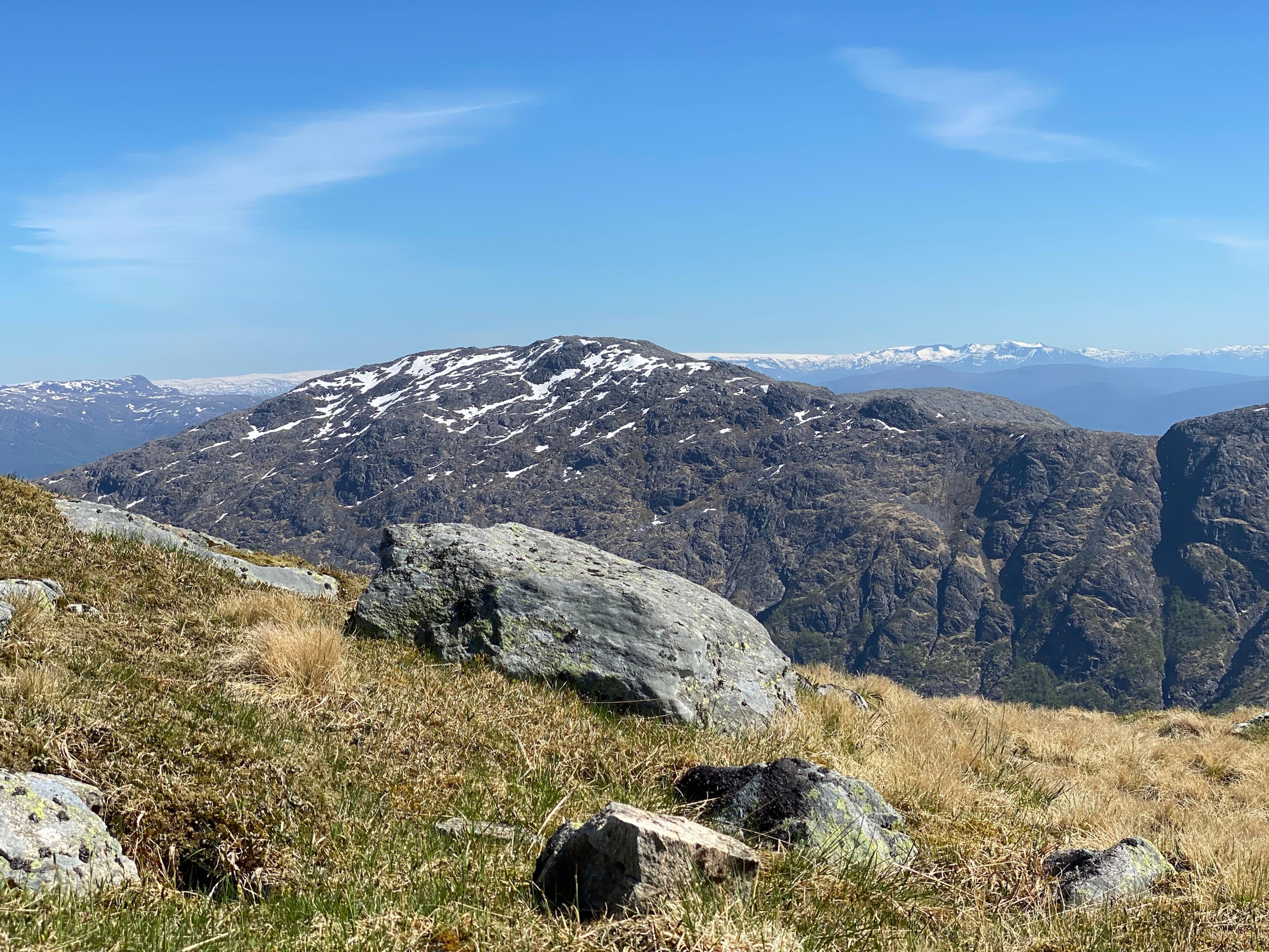 Stor steinblokk i gul fjellgress i forgrunnen, og et bart, avrundet fjell med snøflekker bak. Langt i bakgrunnen ligger snøkledde fjell og en isbre.