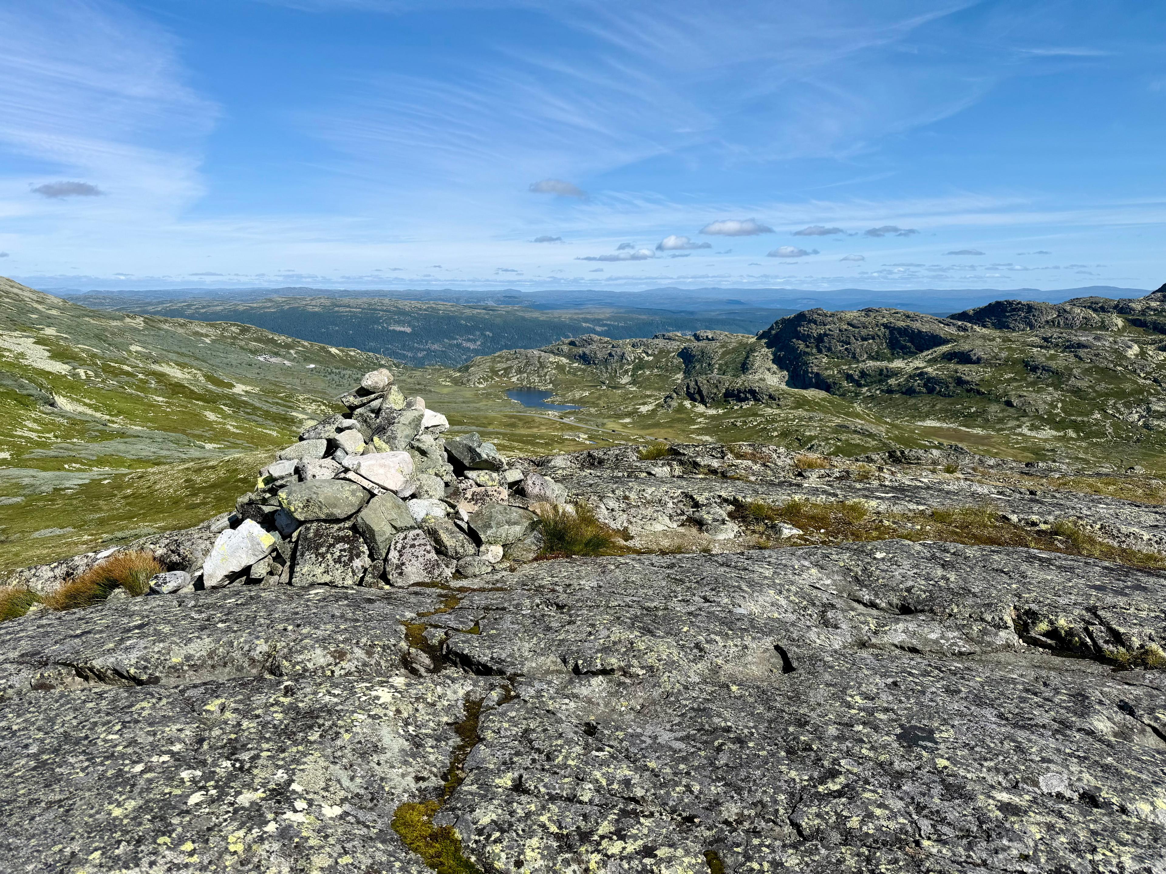 Steinvarde på bart fjell, med vidde, vann og fjell i bakgrunnen under blå himmel.