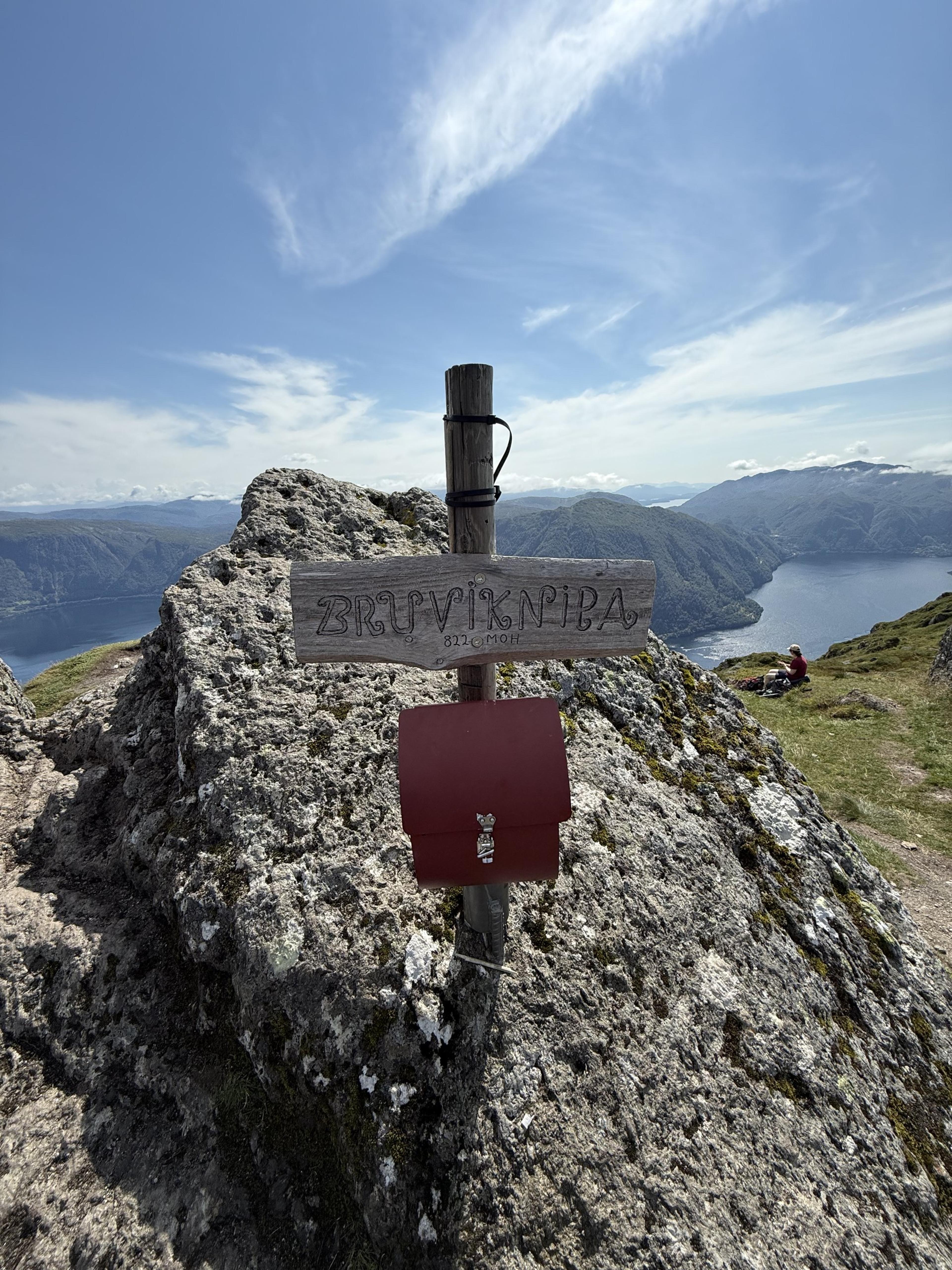 Treskilt med teksten «Bruviknipa 822 moh.» på en trestolpe over en rød postkasse, på en grå bergknaus med fjord og fjell bak.