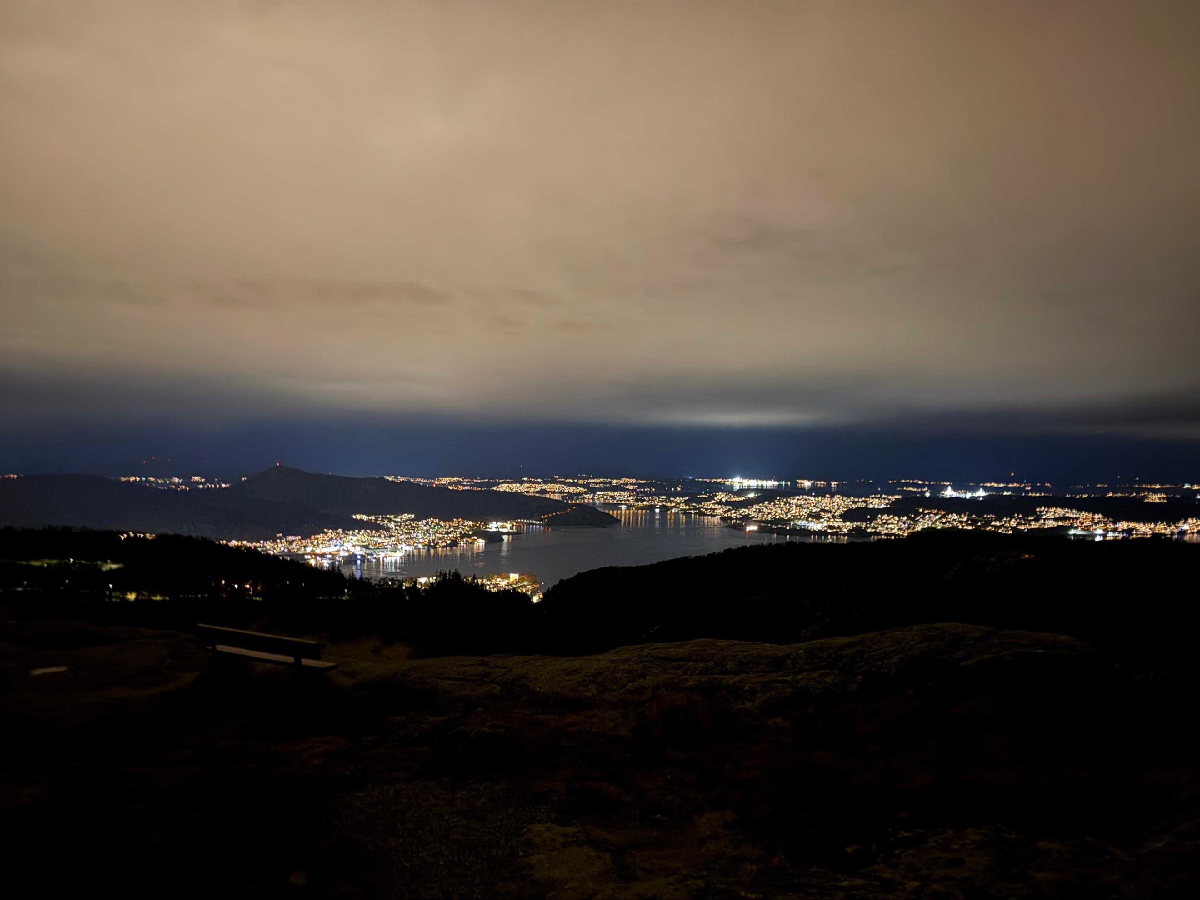 Bylys langs en fjord om kvelden, med mørke åser i forgrunnen og et lavt, lyst skydekke over.