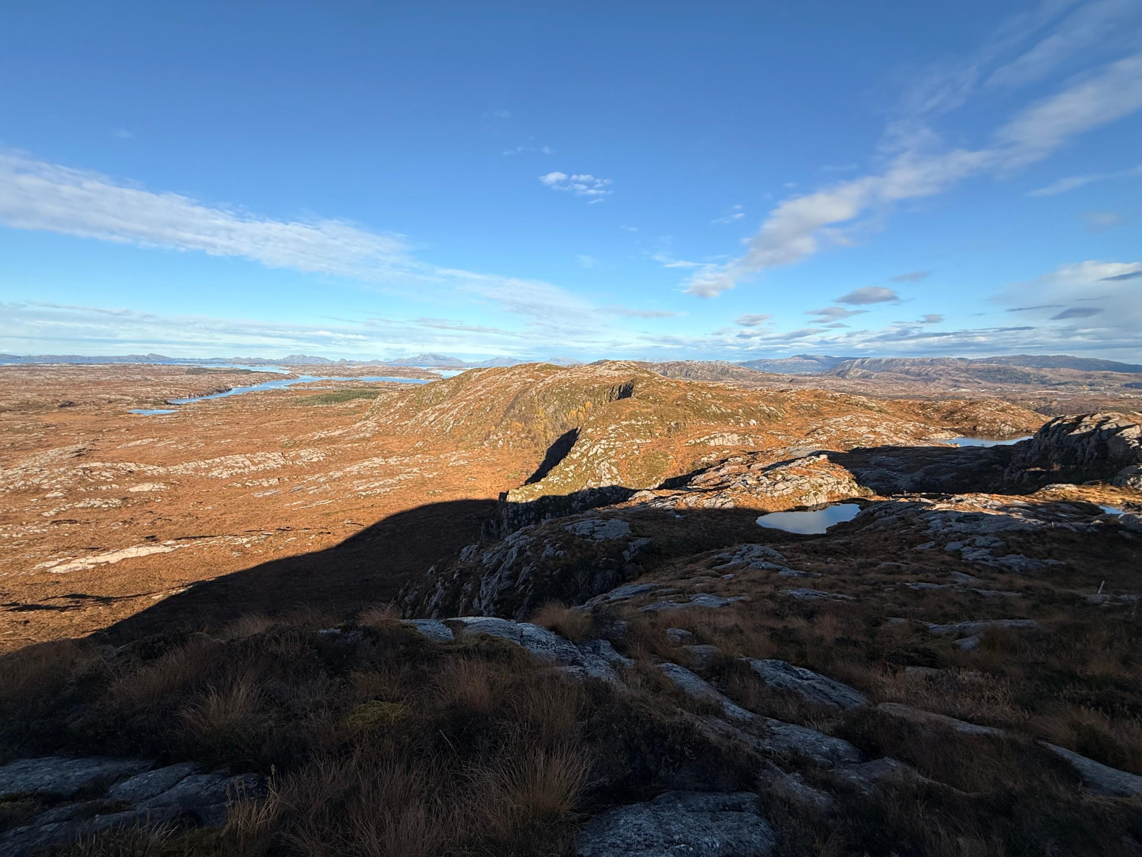 Utsikt fra en lyngkledd topp over brunt lynglandskap med små tjern, en fjordarm og fjell i horisonten. Toppens skygge faller over terrenget i forgrunnen.