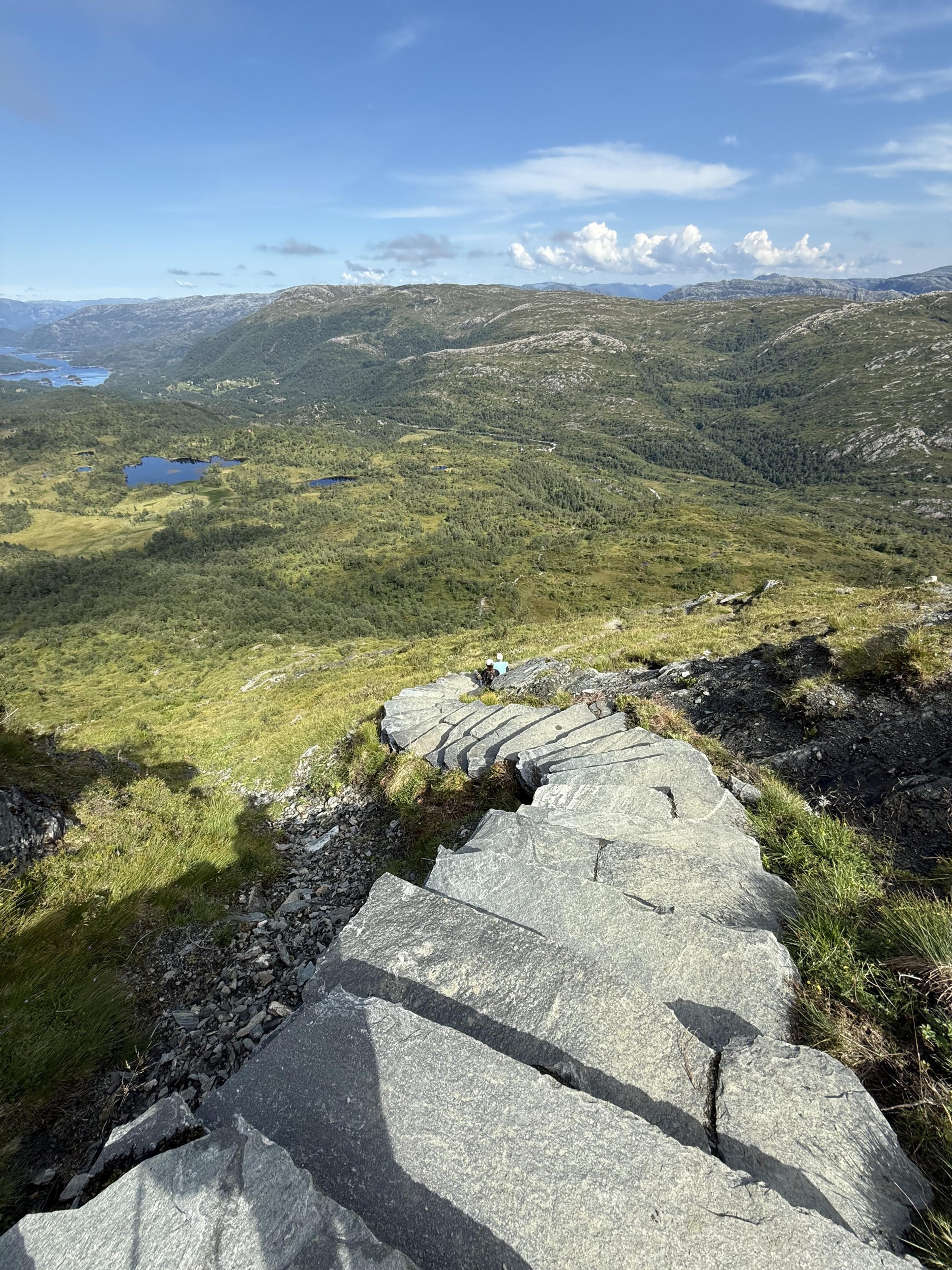 Steinlagte trapper i bratt, gresskledd terreng, med skog, små vann og åser langt nedenfor.
