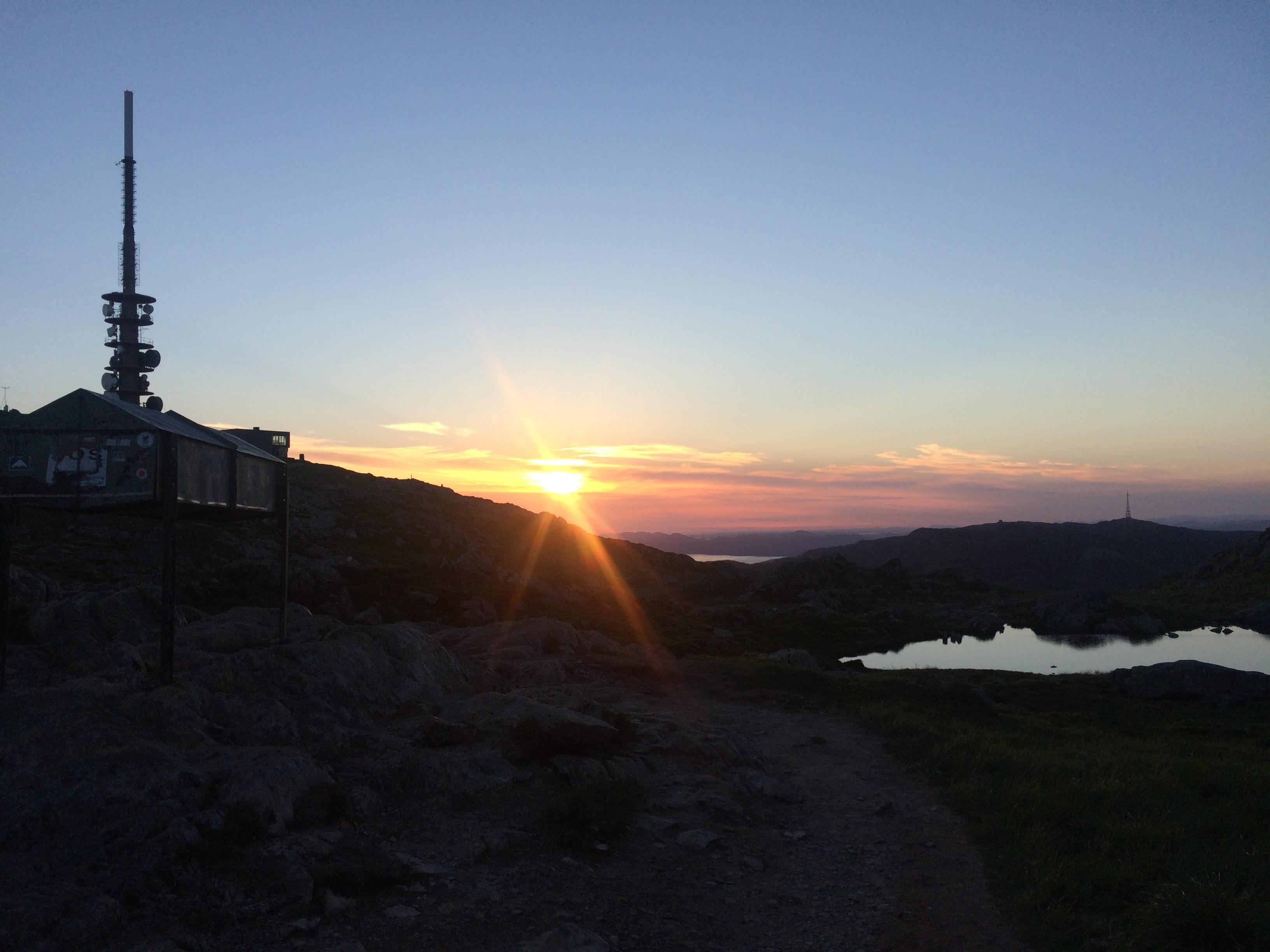 Solnedgang på Ulriken med masta i silhuett og et lite vann som speiler himmelen.