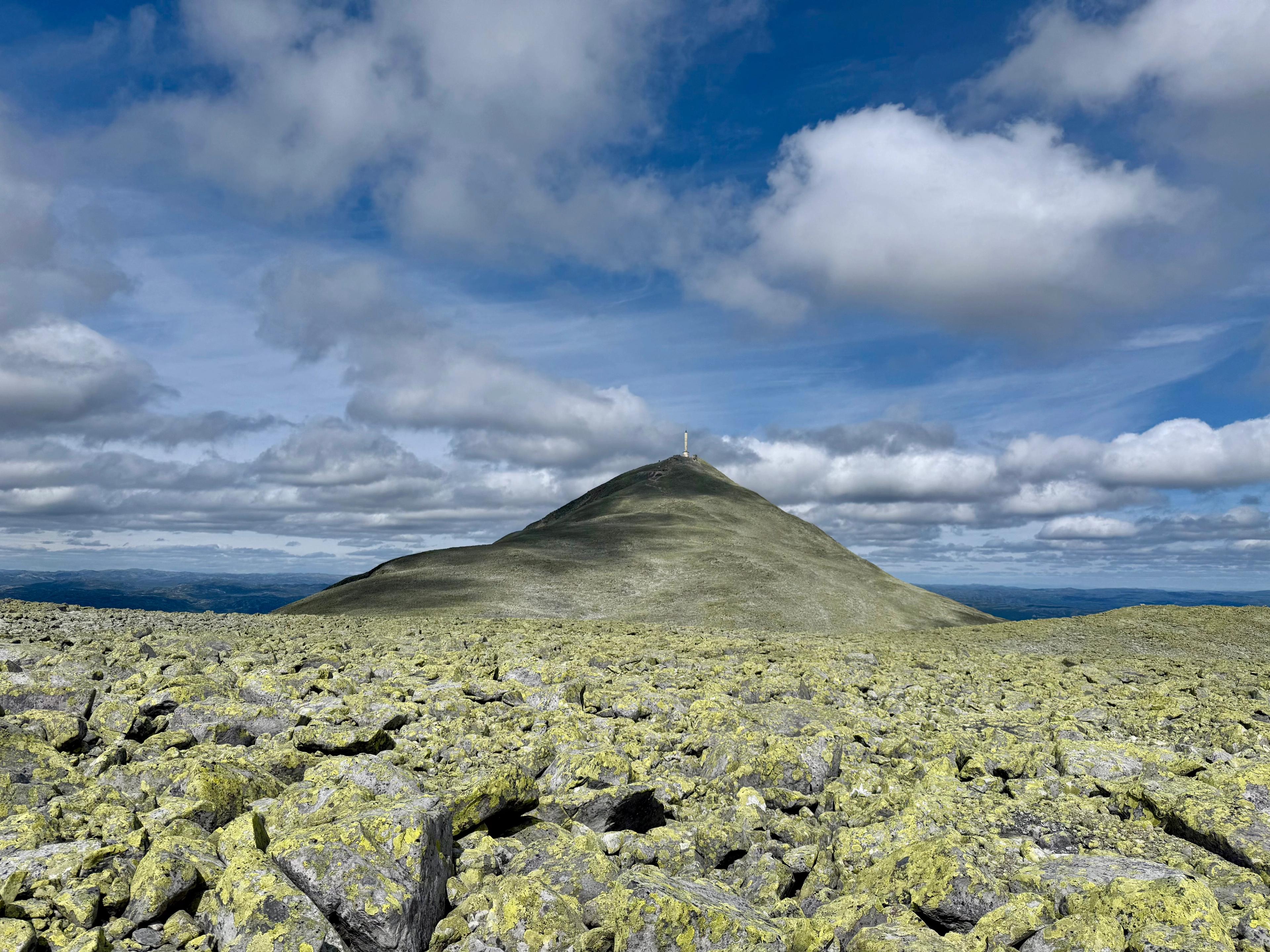Grønn, kjegleformet fjelltopp med et tårn på toppen, sett over en steinur under blå himmel med skyer.