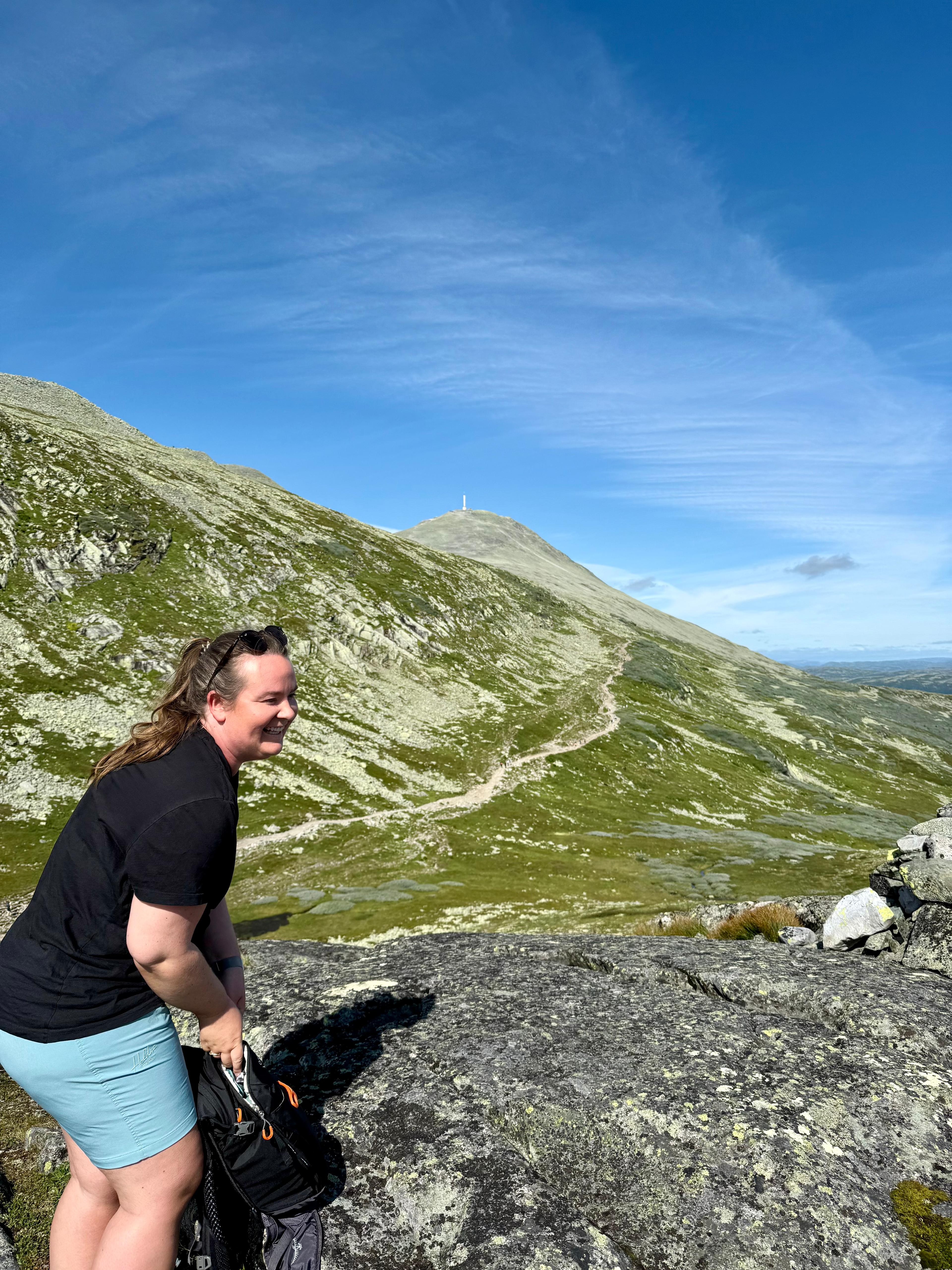 Smilende kvinne ved en stor stein, med Gaustatoppen og stien opp i bakgrunnen.