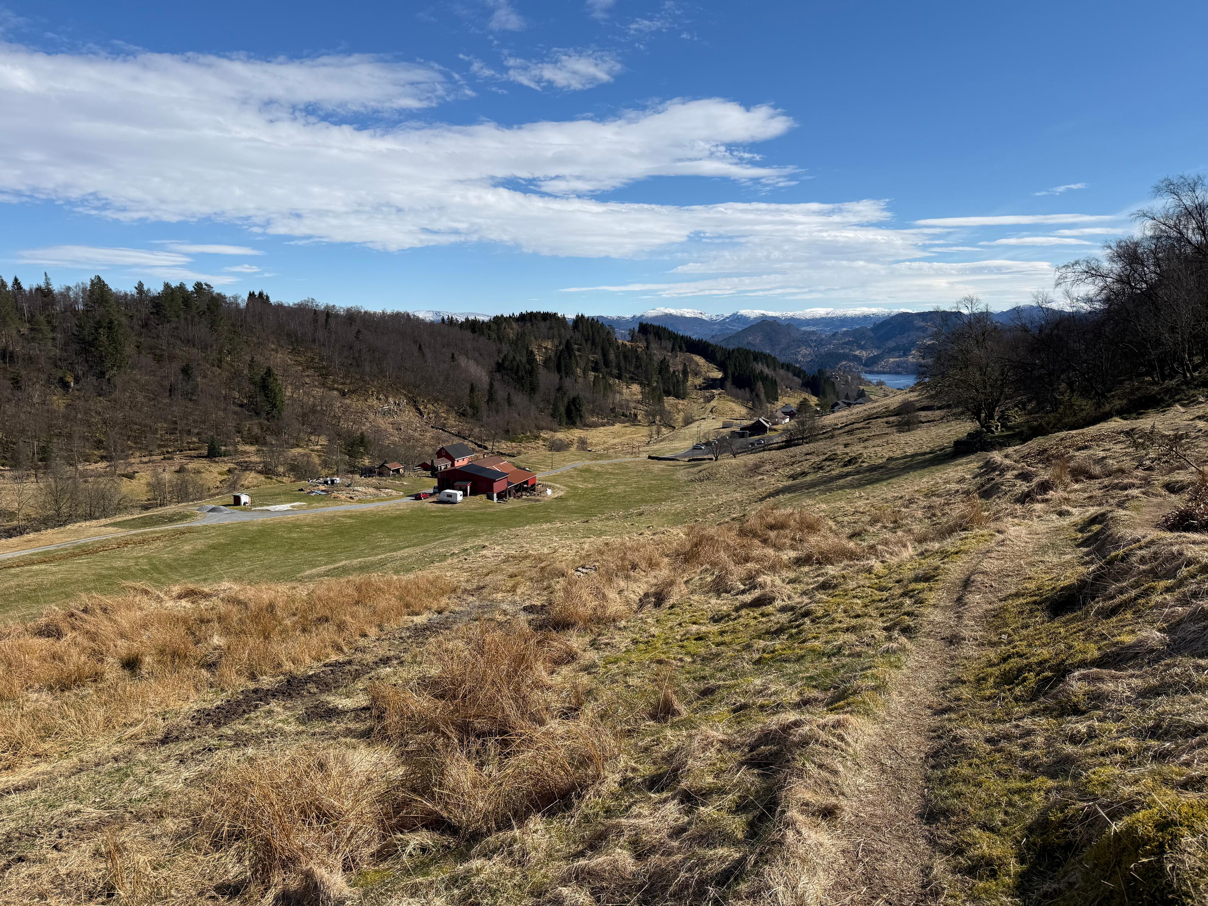 Smal sti gjennom gulnet gress ned mot en rød gård, med skog og snødekte fjell i bakgrunnen.