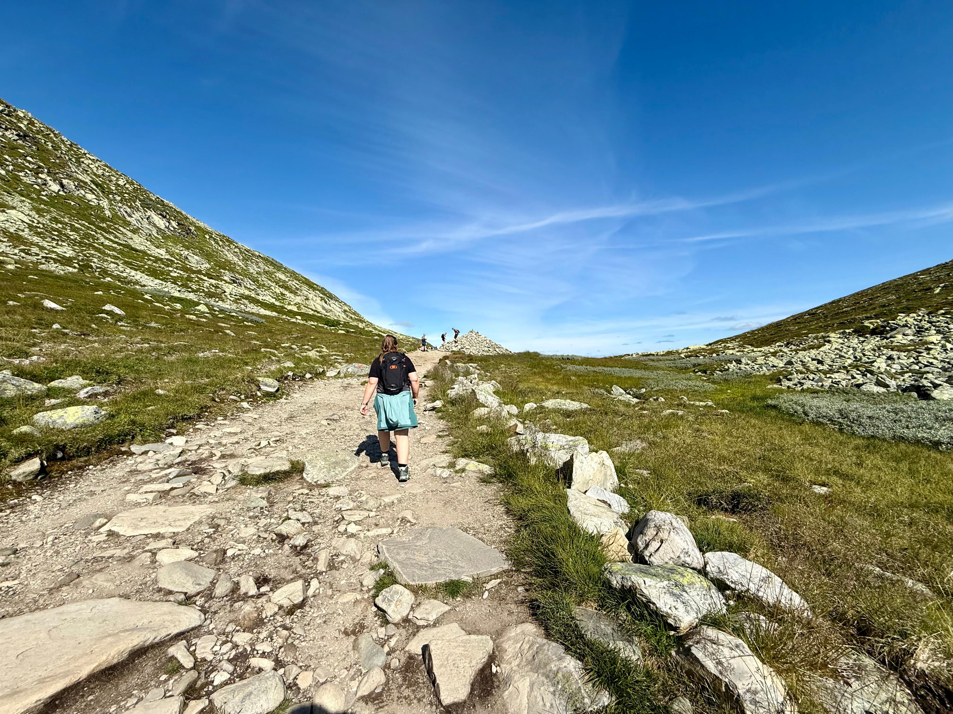 Kvinne går på bred steinsti oppover mellom grønne fjellsider, med folk lenger fremme.