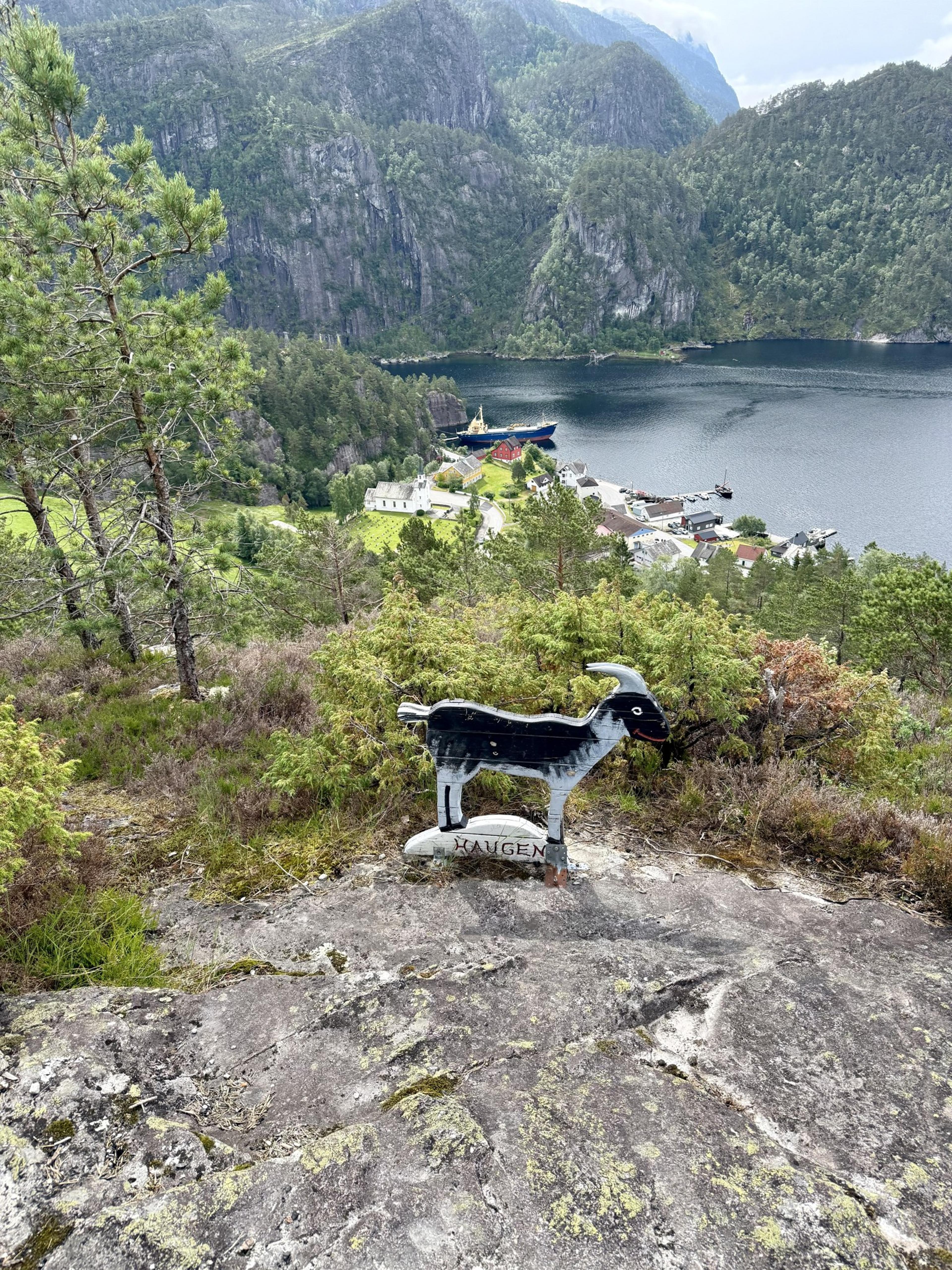 Svart og hvit geitefigur i tre på et berg, med fjorden og bygden langt nede bak.