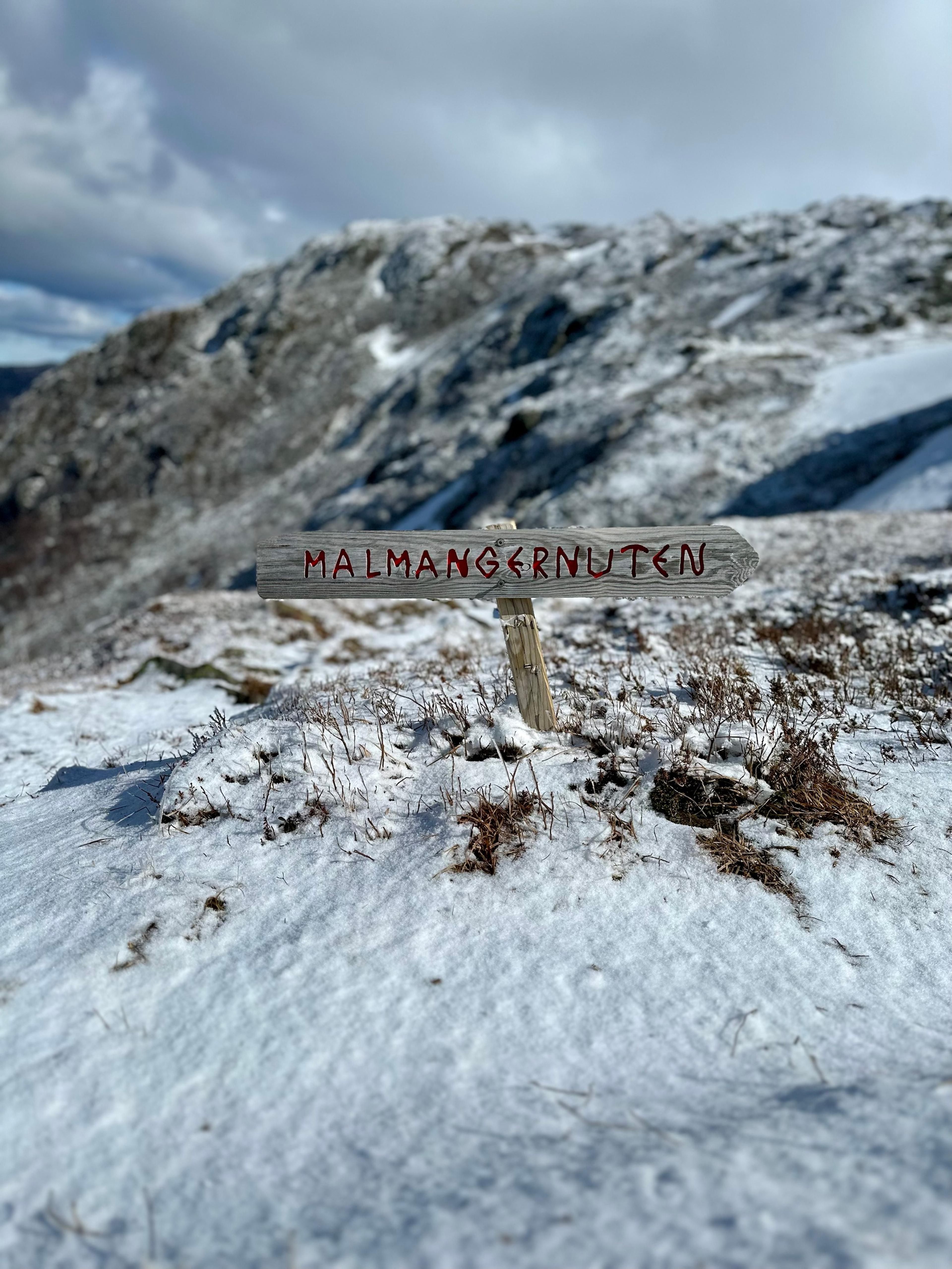 Håndlaget treskilt med rød tekst «Malmangernuten» og pil mot høyre, i snø og lyng.