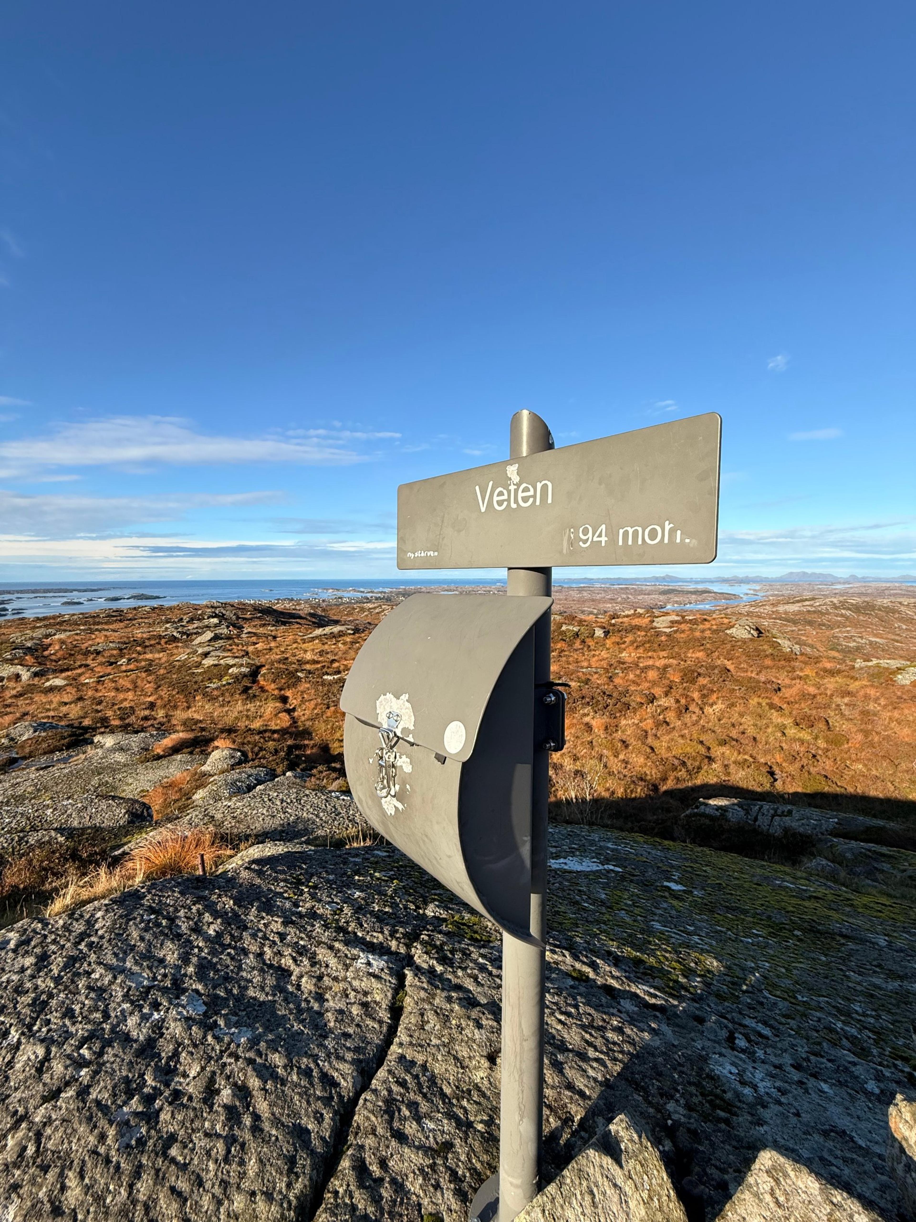 Grått skilt med teksten «Veten 194 moh.» og en postkasse på en stolpe, på et svaberg med lyngmark og havet bak.