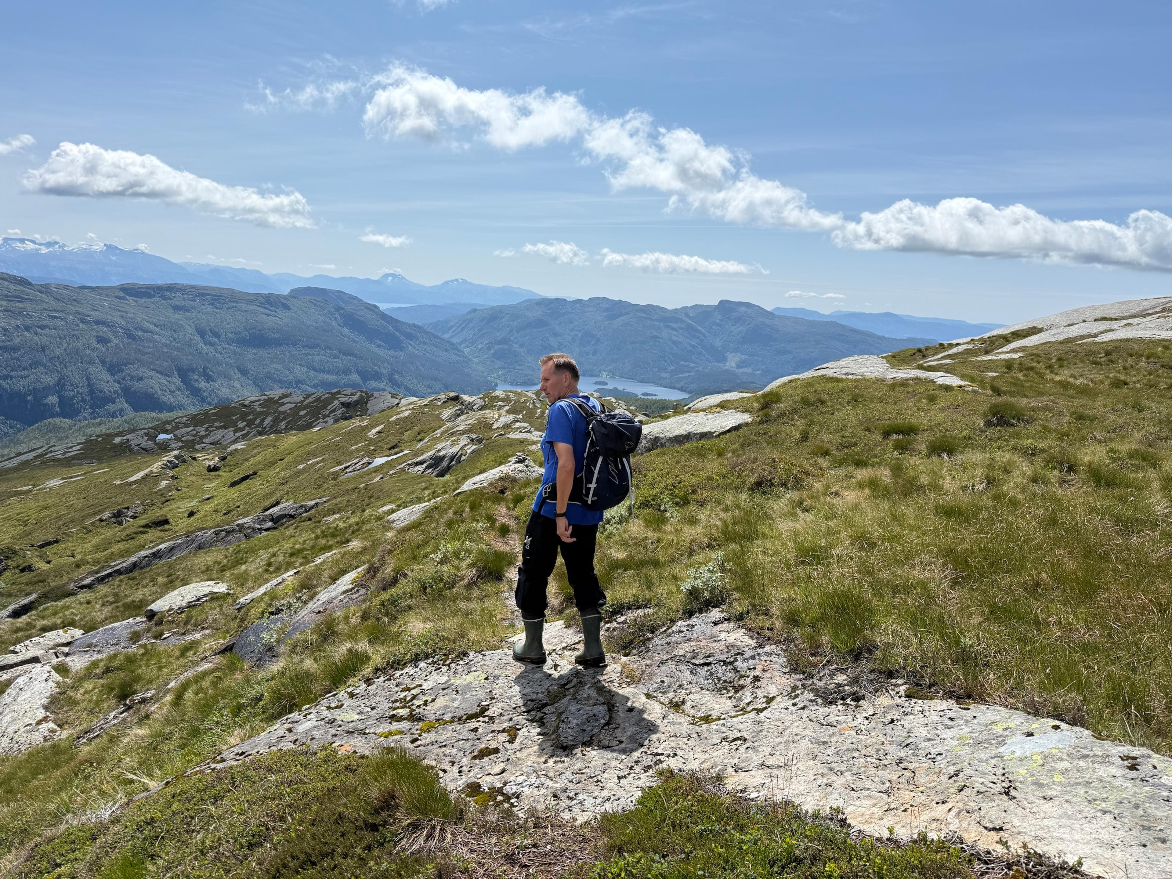 En turgåer i blå genser sitter på et svaberg ved en bratt fjellkant og ser ut over en grønn dal med flere vann på rad ned mot en fjord.