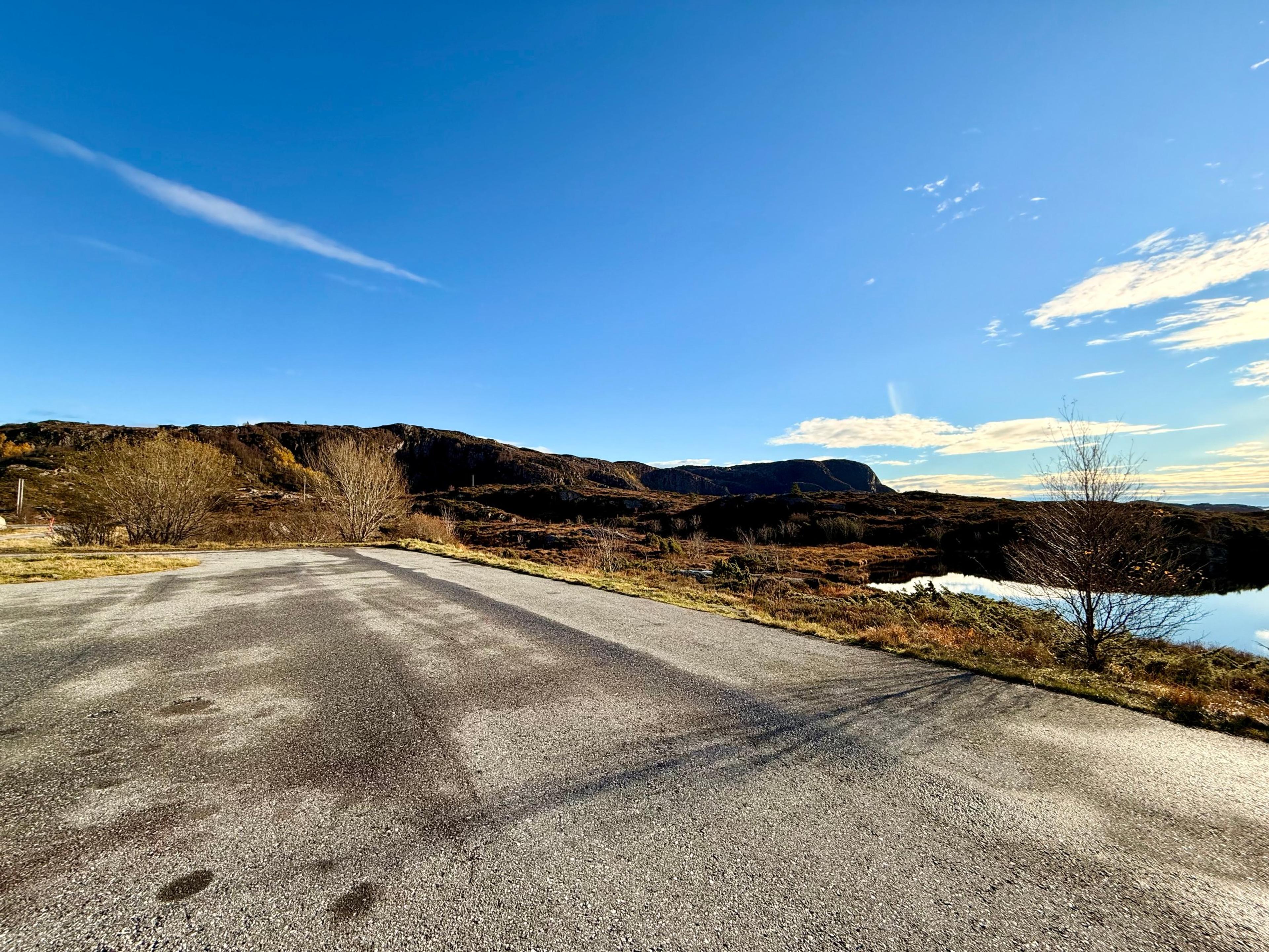 Tom asfaltert parkeringsplass ved et lite tjern, med lyng og bjørk rundt og en bratt fjellrygg bak under blå himmel.