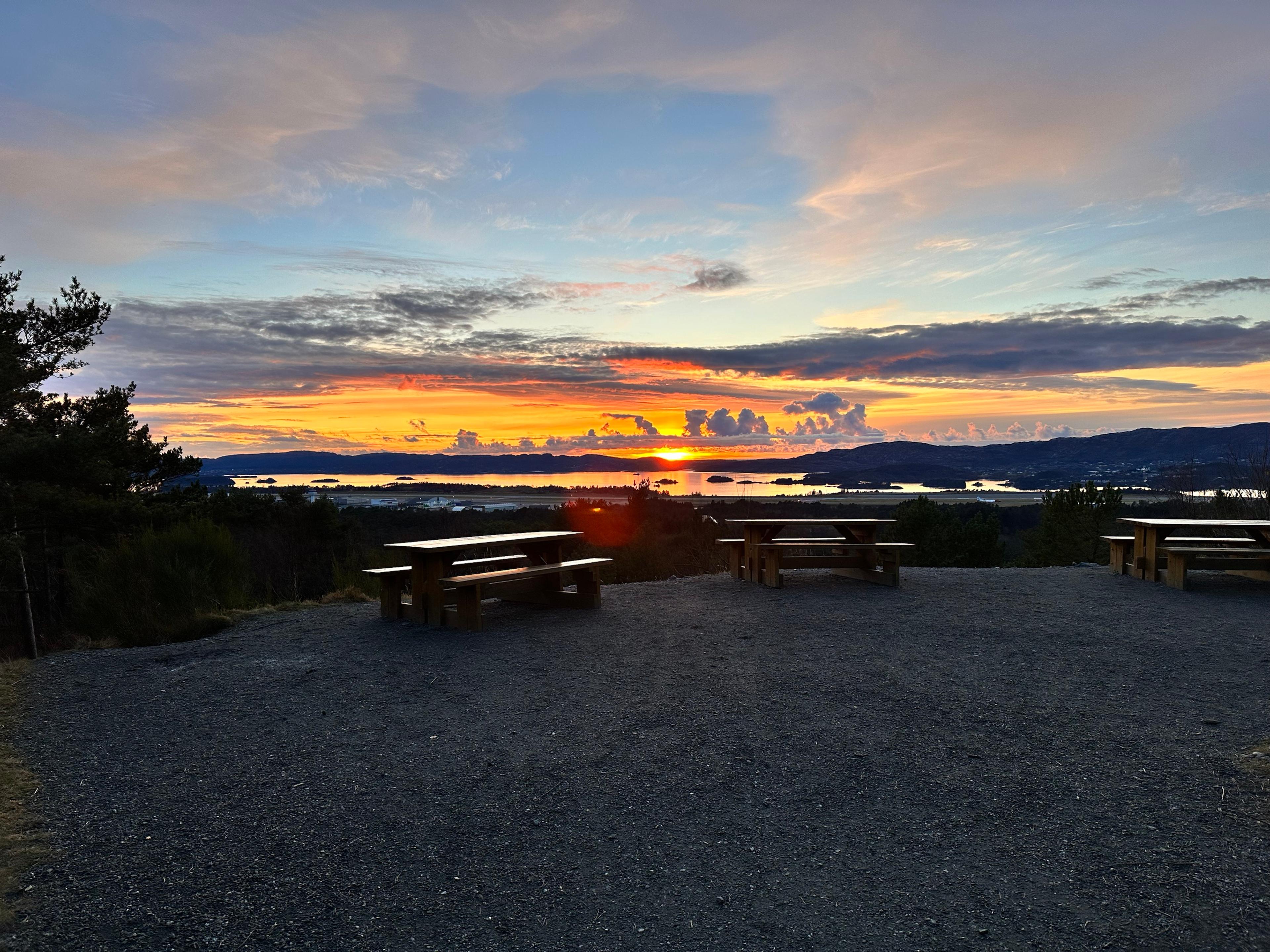 Solnedgang over sjøen og holmene sett fra Storrinden, med tre benker på en gruslagt plass og Flesland nedenfor.