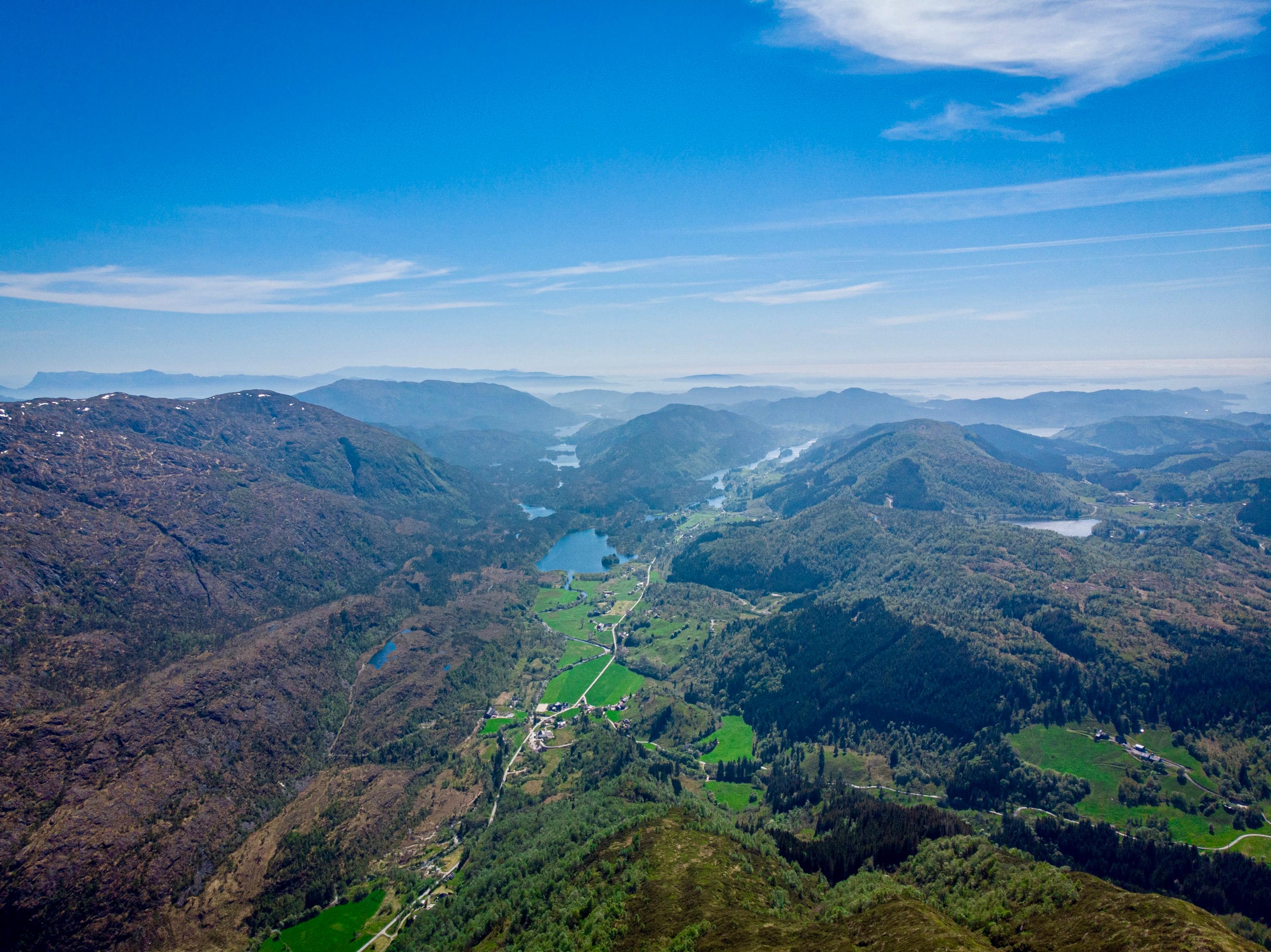 Dronebilde over en lang dal med jorder, gårder og en vei, et vann med en liten holme, og skogkledde fjell på begge sider ut mot kysten.