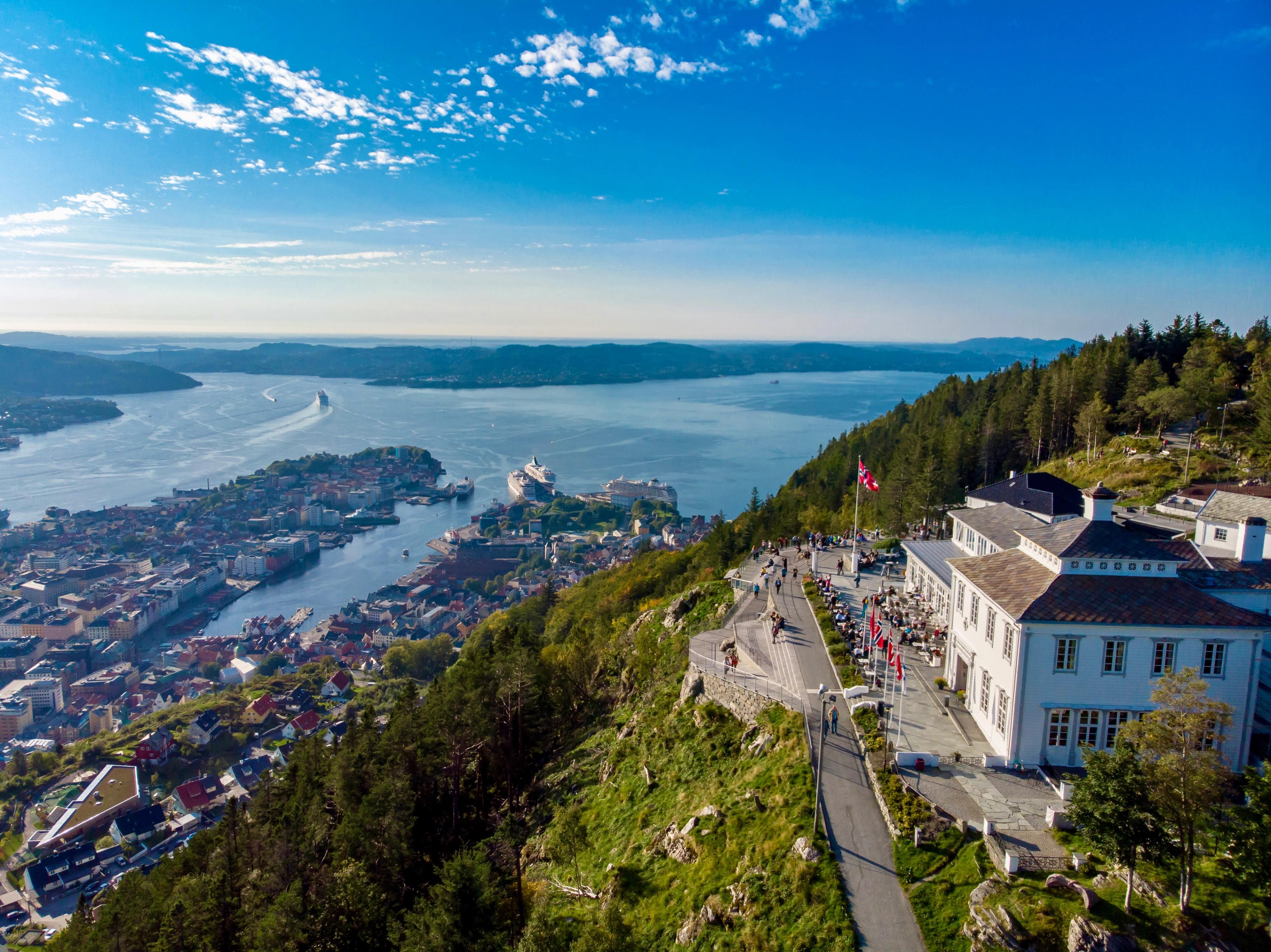 Dronebilde av Fløyen i solskinn. Den hvite trebygningen til Fløien Folkerestaurant ligger til høyre med uteservering og norske flagg, og nedenfor til venstre ligger Bergen sentrum og Byfjorden med cruiseskip ved kai.