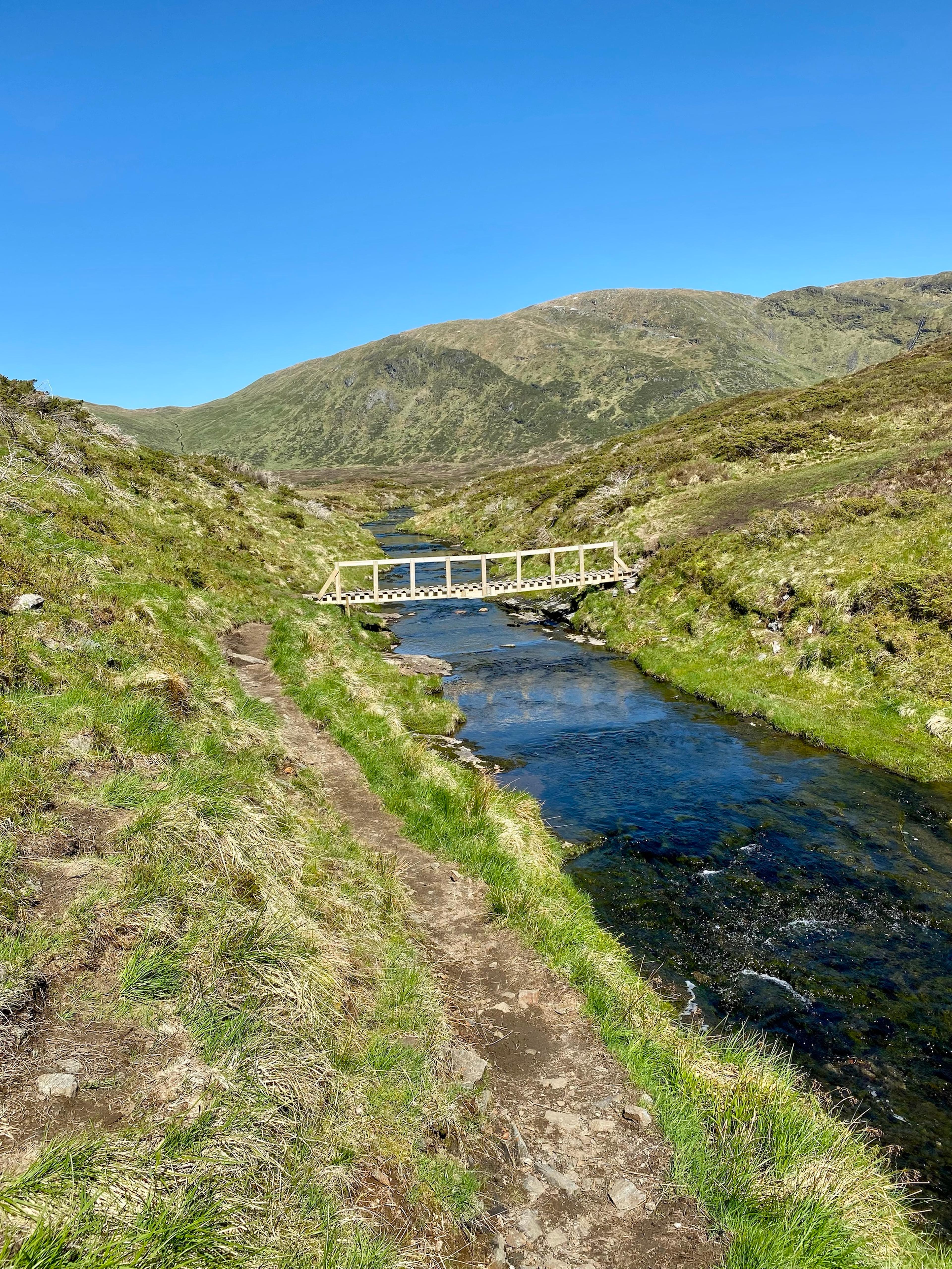 Sti langs en elv i en gressgrodd dal, med en trebro over elven og en avrundet fjellside i bakgrunnen.