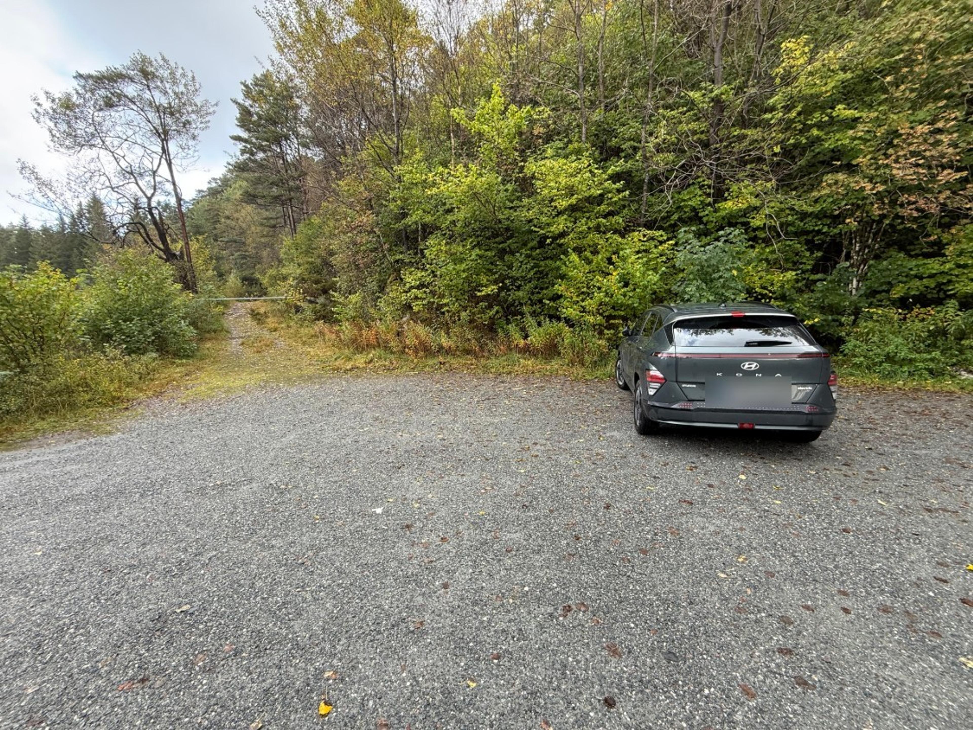 Grusplass med en parkert bil foran løvskog. Til venstre går en gresskledd grusvei inn i skogen, stengt med en bom.