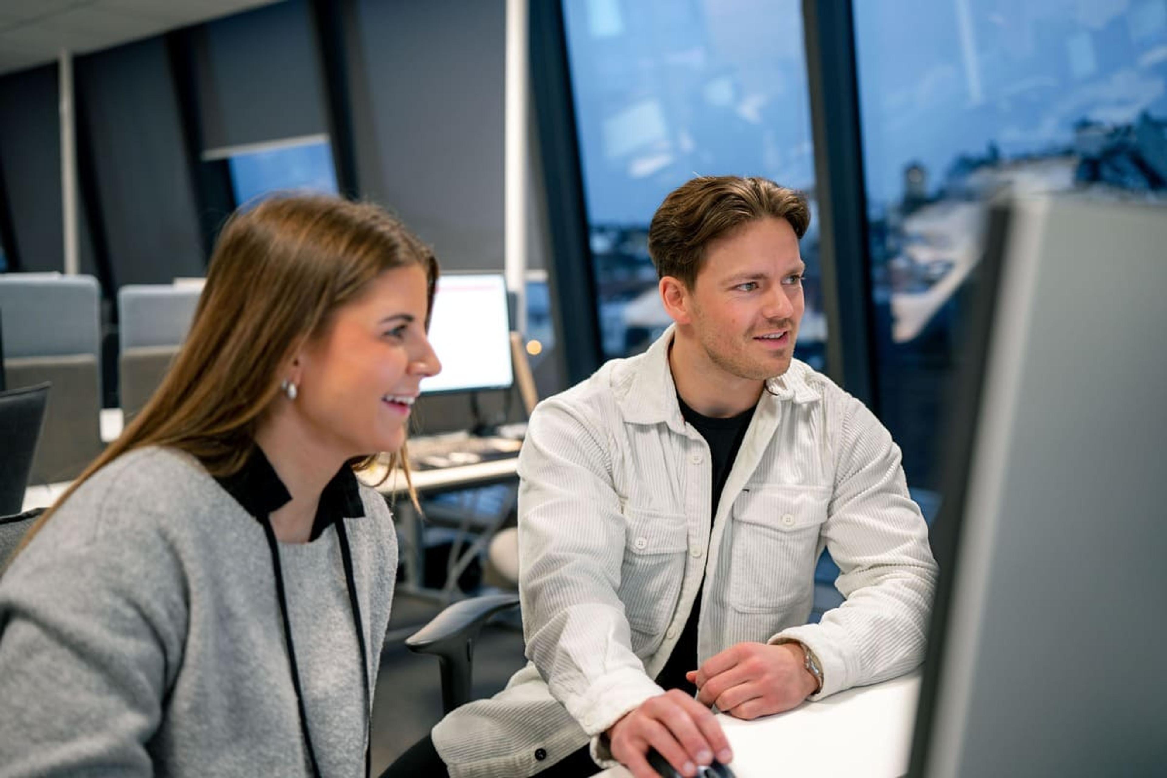 Bilde av dame som smiler og mann som ser på PC skjerm og sitter ved en pult