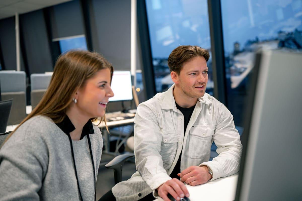 Bilde av dame som smiler og mann som ser på PC skjerm og sitter ved en pult