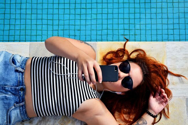 Dame med solbriller som ligger på bassengkanten og ser på mobilen sin