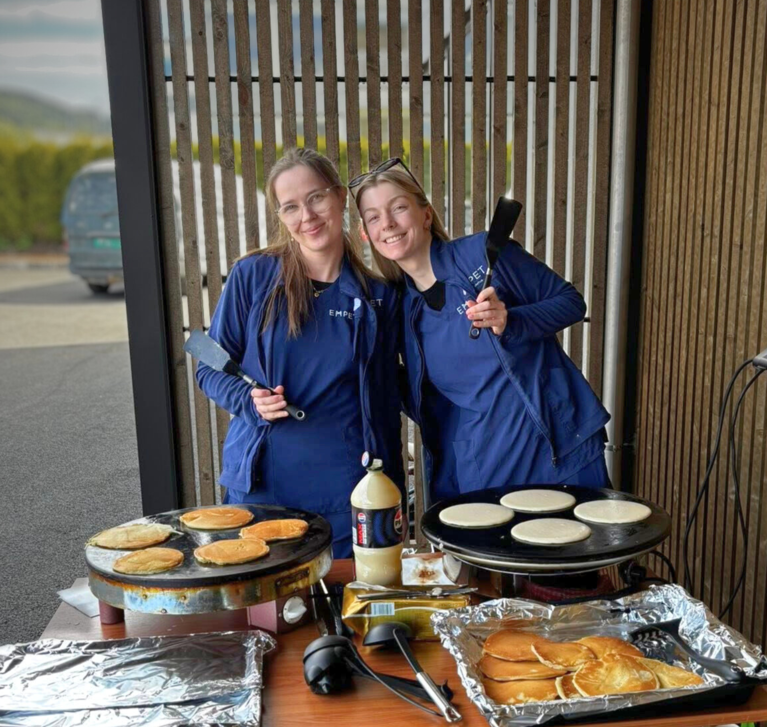 To ansatte i blå EMPET-uniformer står smilende ved et bord ute og steker pannekaker, med tallerkener fulle av ferdige pannekaker foran seg.