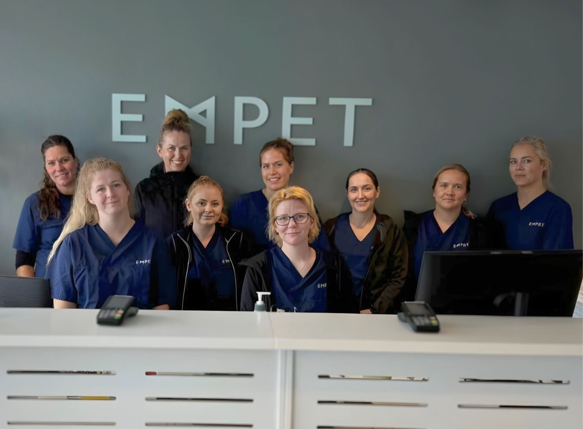 Ansatte ved EMPET Askim står samlet bak resepsjonsdisken og smiler foran EMPET-logoen.
