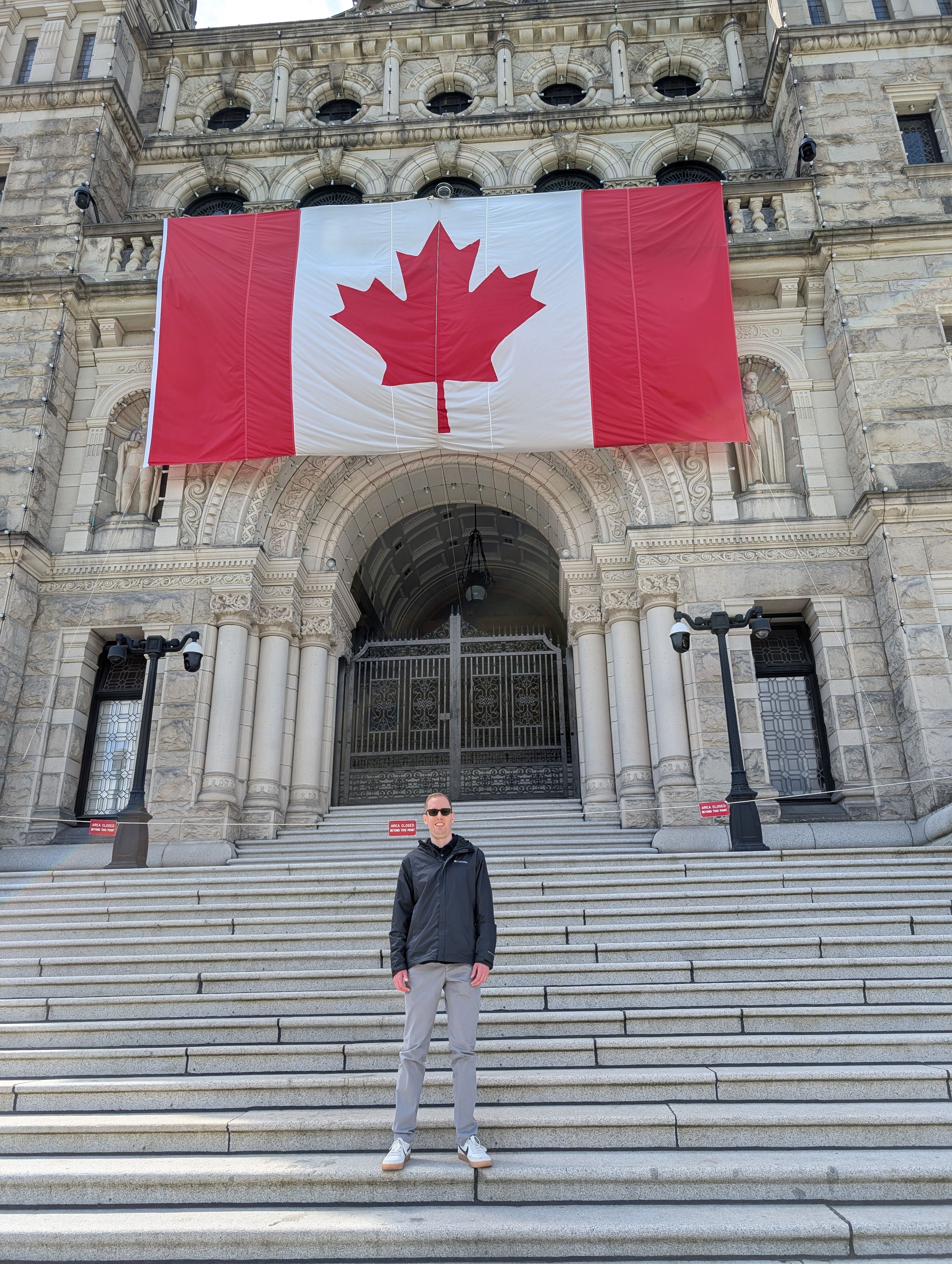 Lucas Silbernagel standing in front of the British Columbia Legislature, with a large Canadian flag on the front of the building