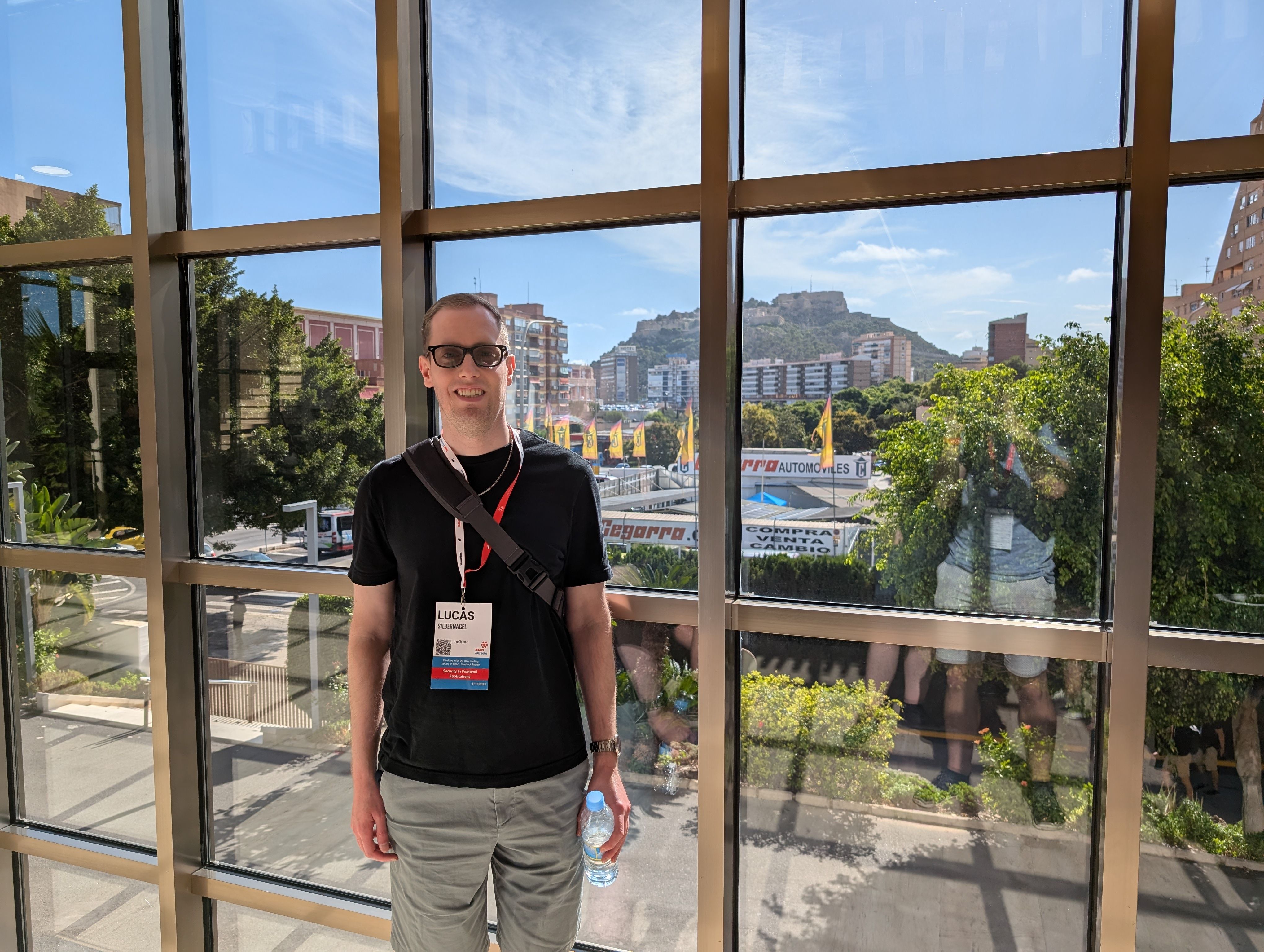 Lucas Silbernagel wearing glasses and a badge for the React Alicante conference, standing in front of a window inside the conference venue, with a sunny view of the Santa Bàrbara castle in the background.