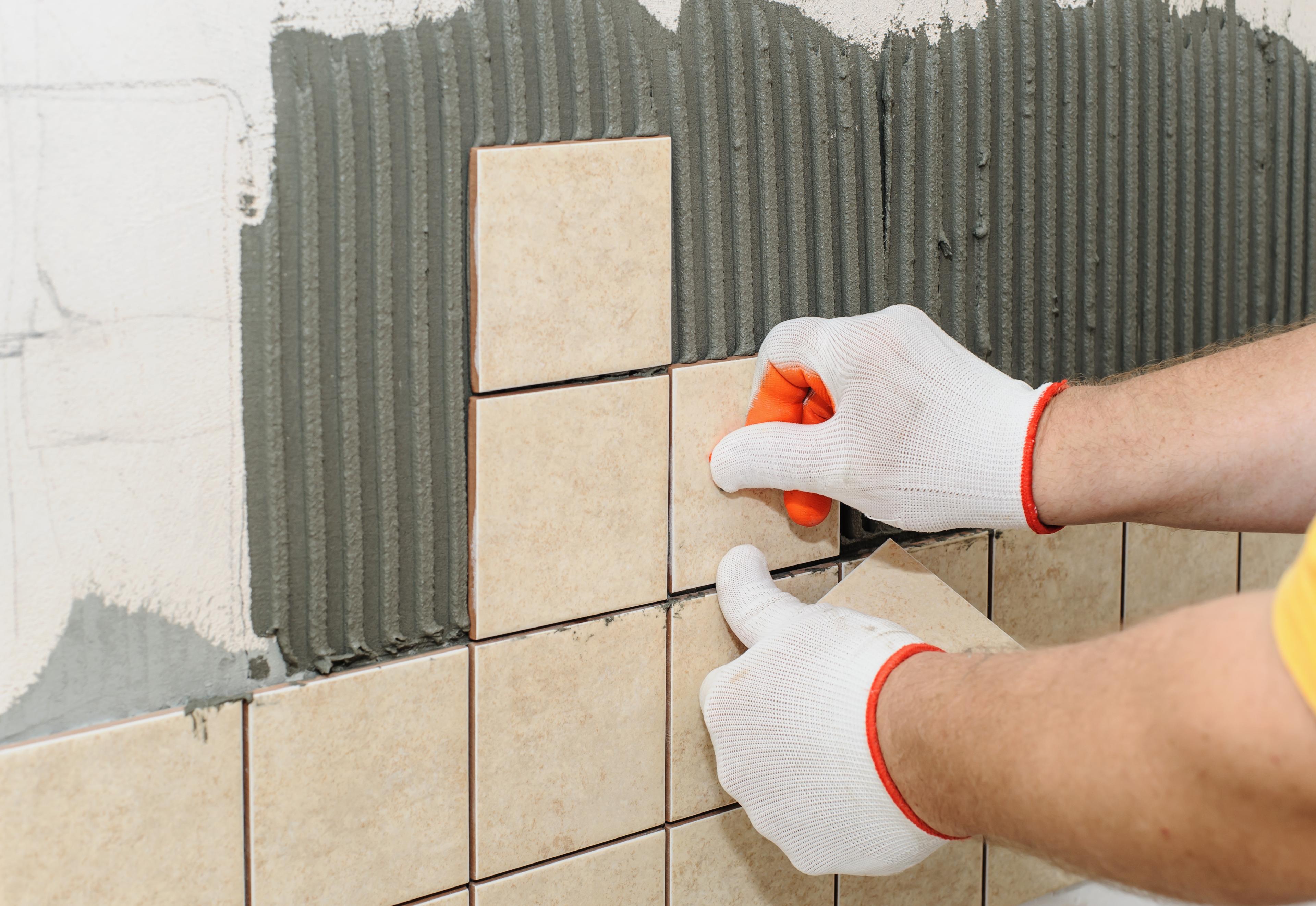 Handskbeklädda händer som sätter upp beige kakel på en vägg med fästmassa