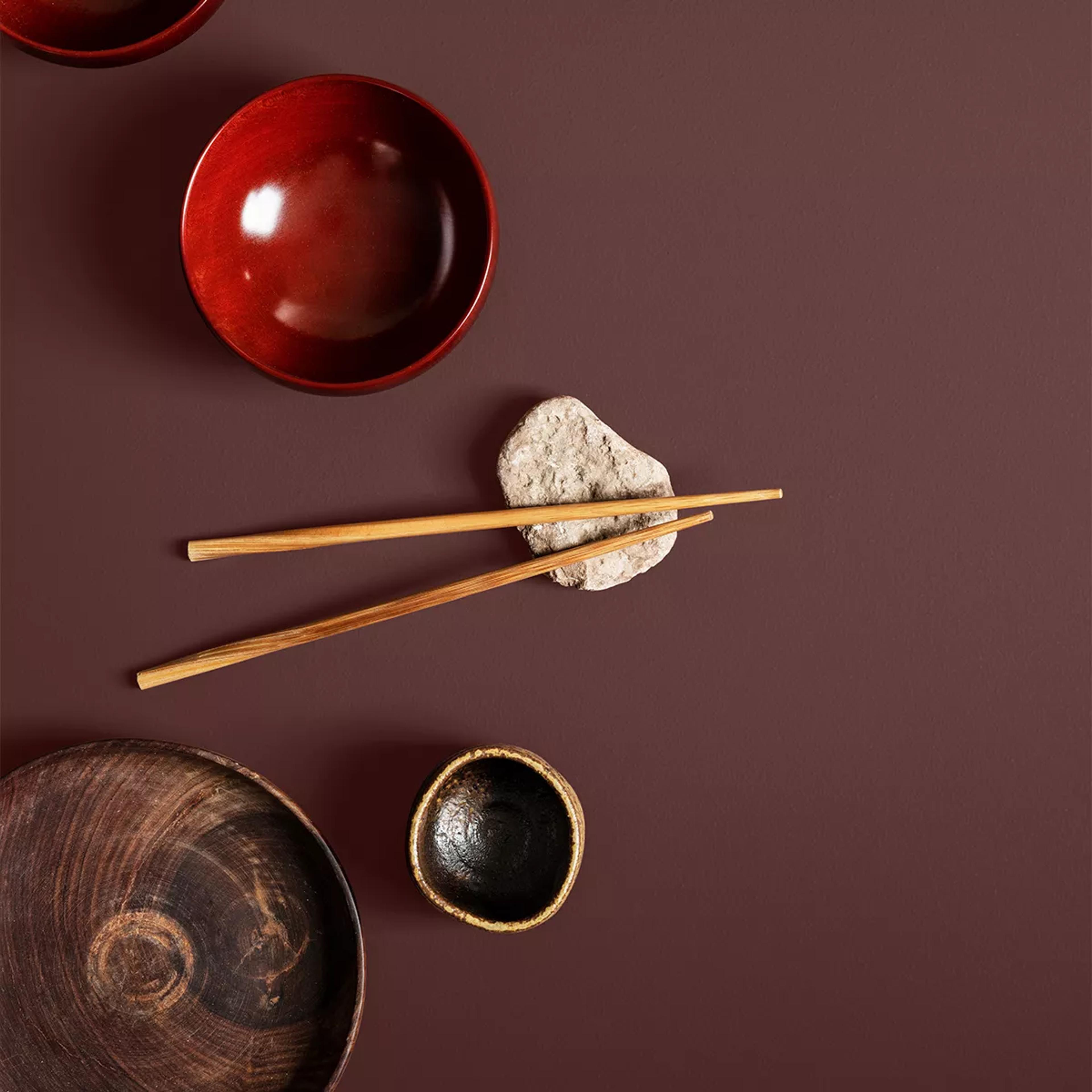 Red bowl, chopsticks on a stone, wooden plate, and small dark bowl on a maroon surface.