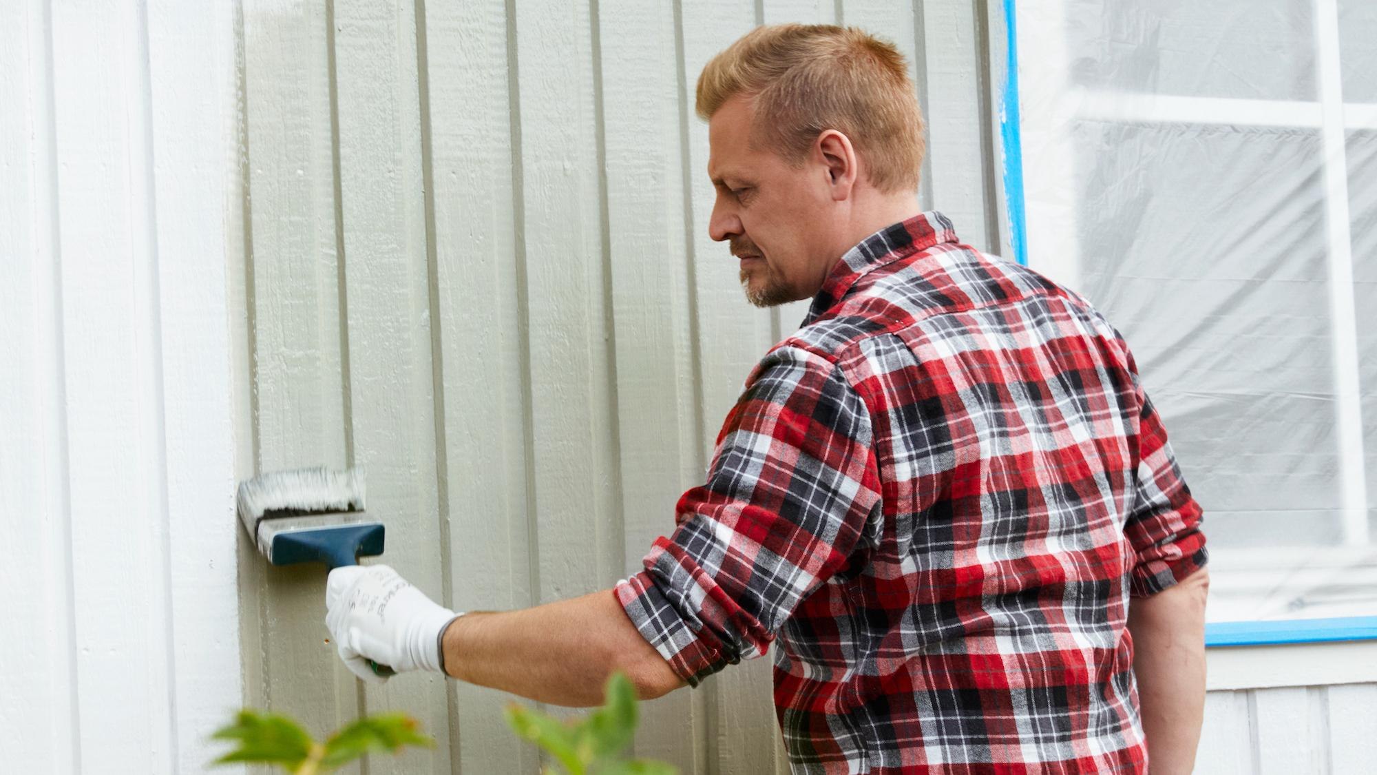 A man in a plaid shirt and gloves paints a house exterior with a paintbrush.