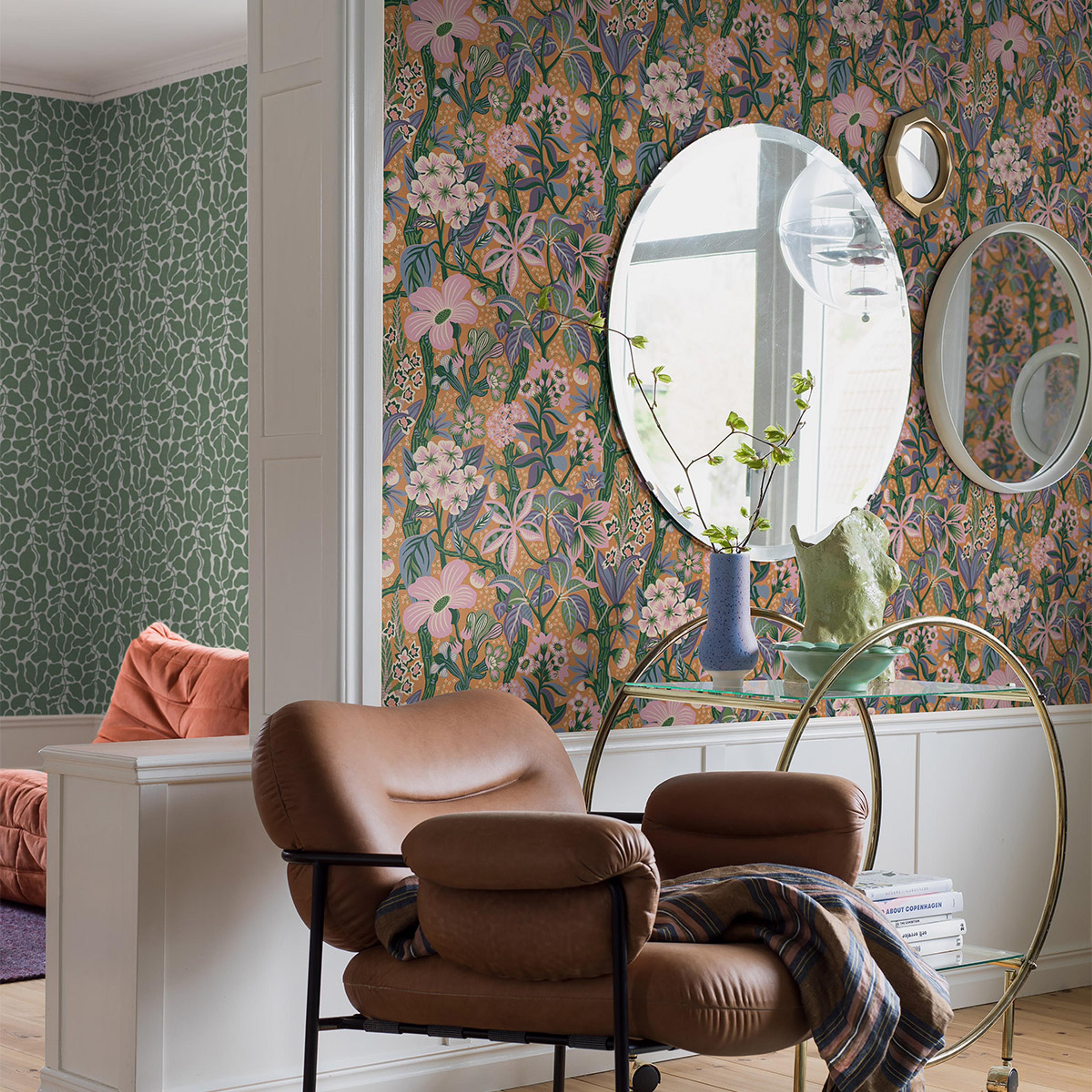 A cozy room with a brown leather armchair, a gold and glass bar cart, and mirrors against a vibrant floral wallpaper, with a green patterned wallpaper visible in an adjacent room.