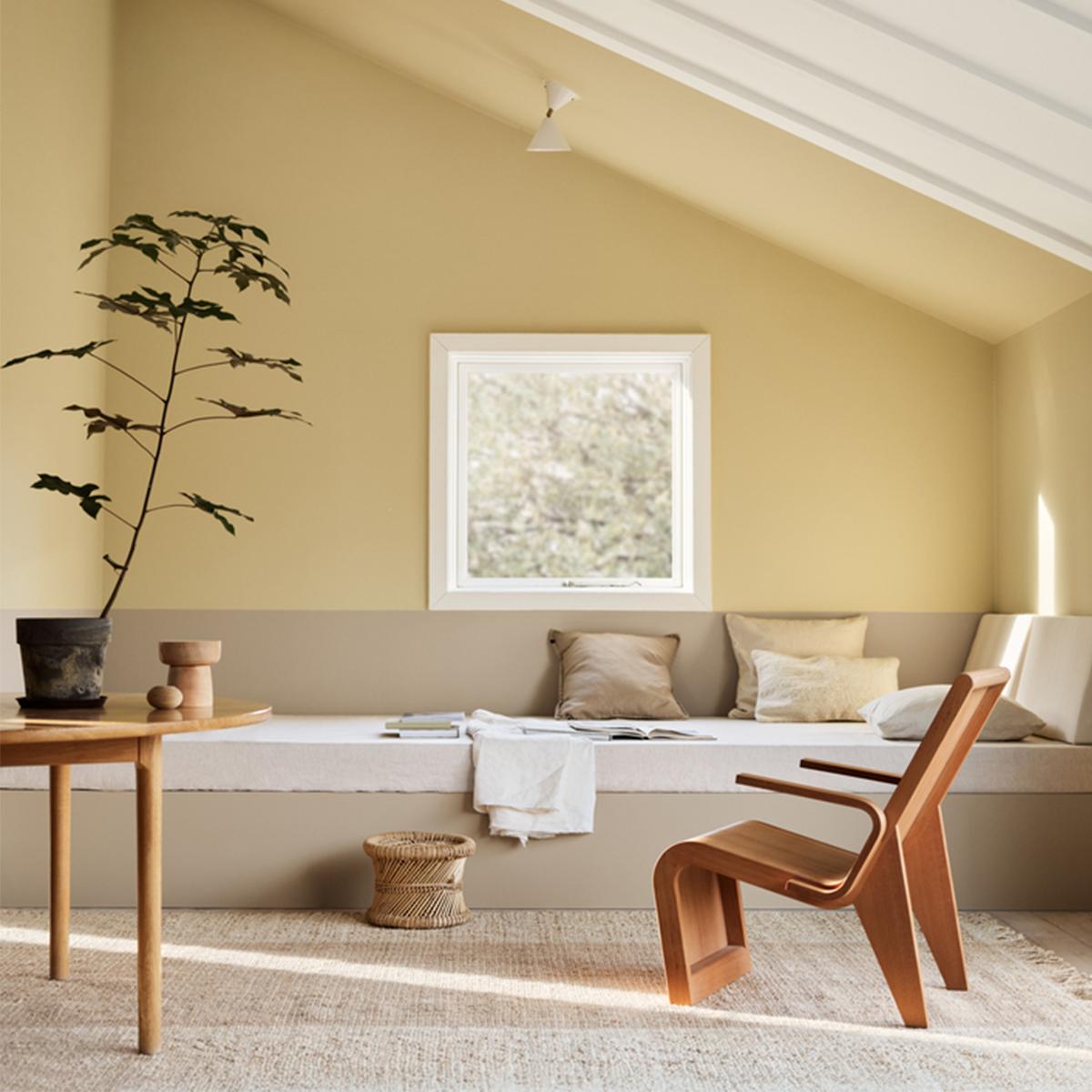 A bright room with pale yellow and beige two-tone walls, featuring a built-in bench under a window, a round wooden table with a plant, and a modern wooden chair.