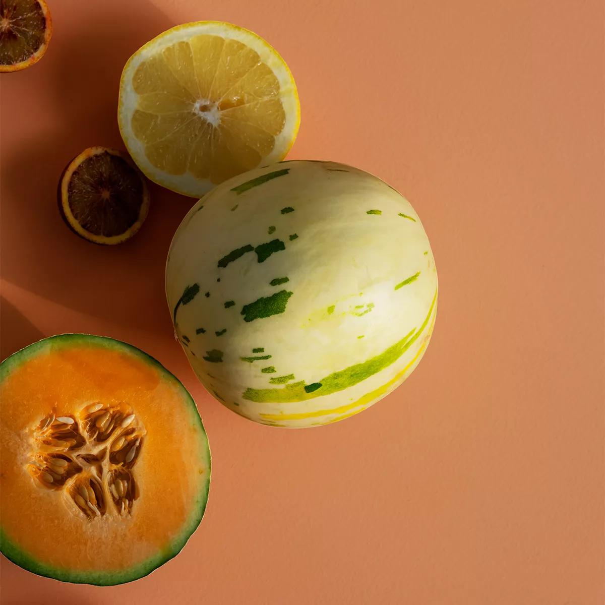 A half lemon, a striped melon, and a half cantaloupe on a peach background.