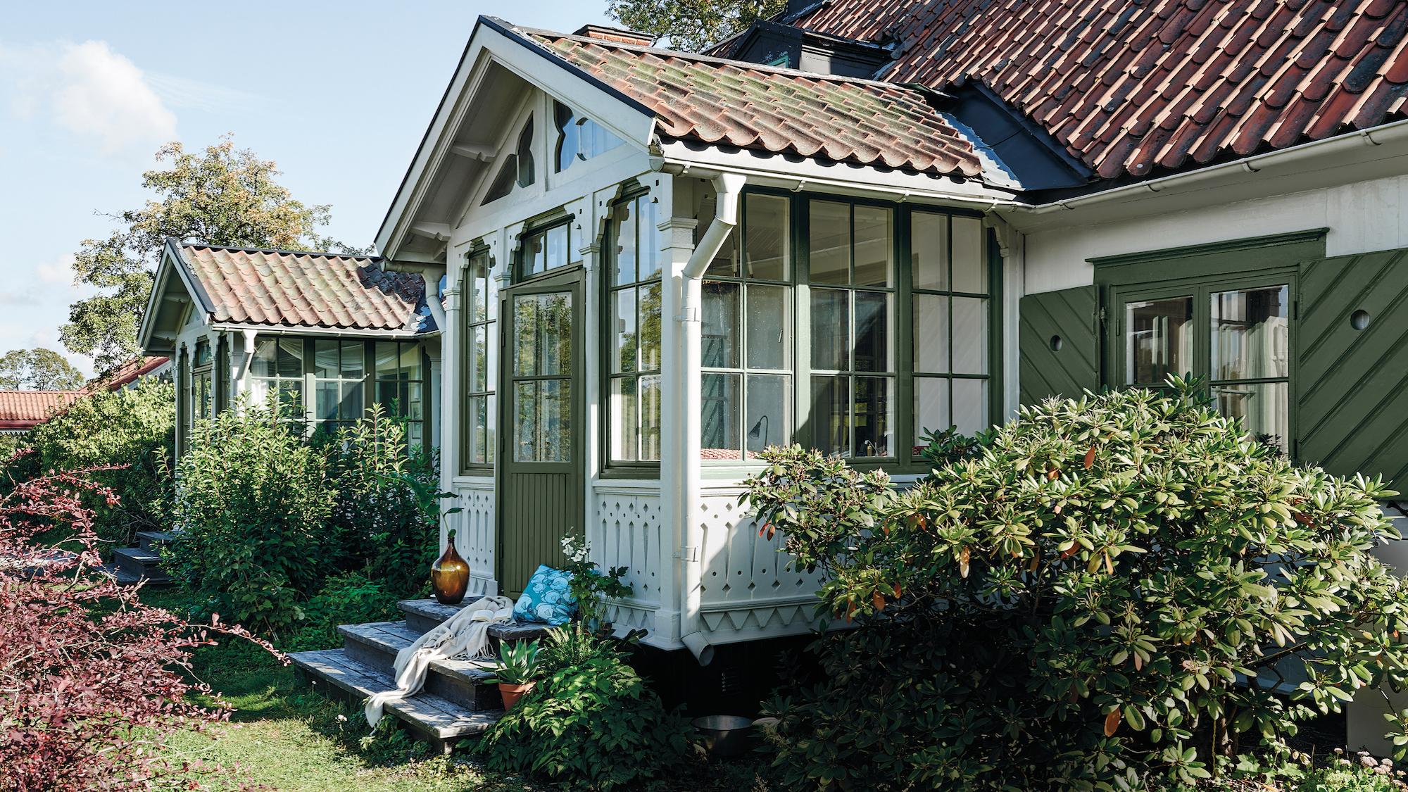 A white house with dark green trim and a red tile roof, featuring ornate gabled porches and surrounded by a lush garden.