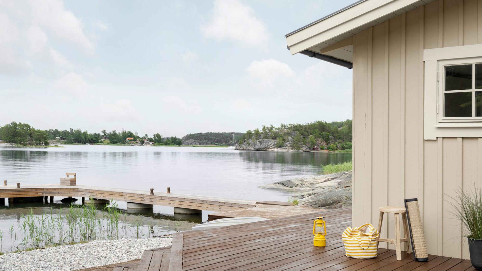 A beige cabin's wooden deck extends to a pier over a calm lake with rocky, forested islands.