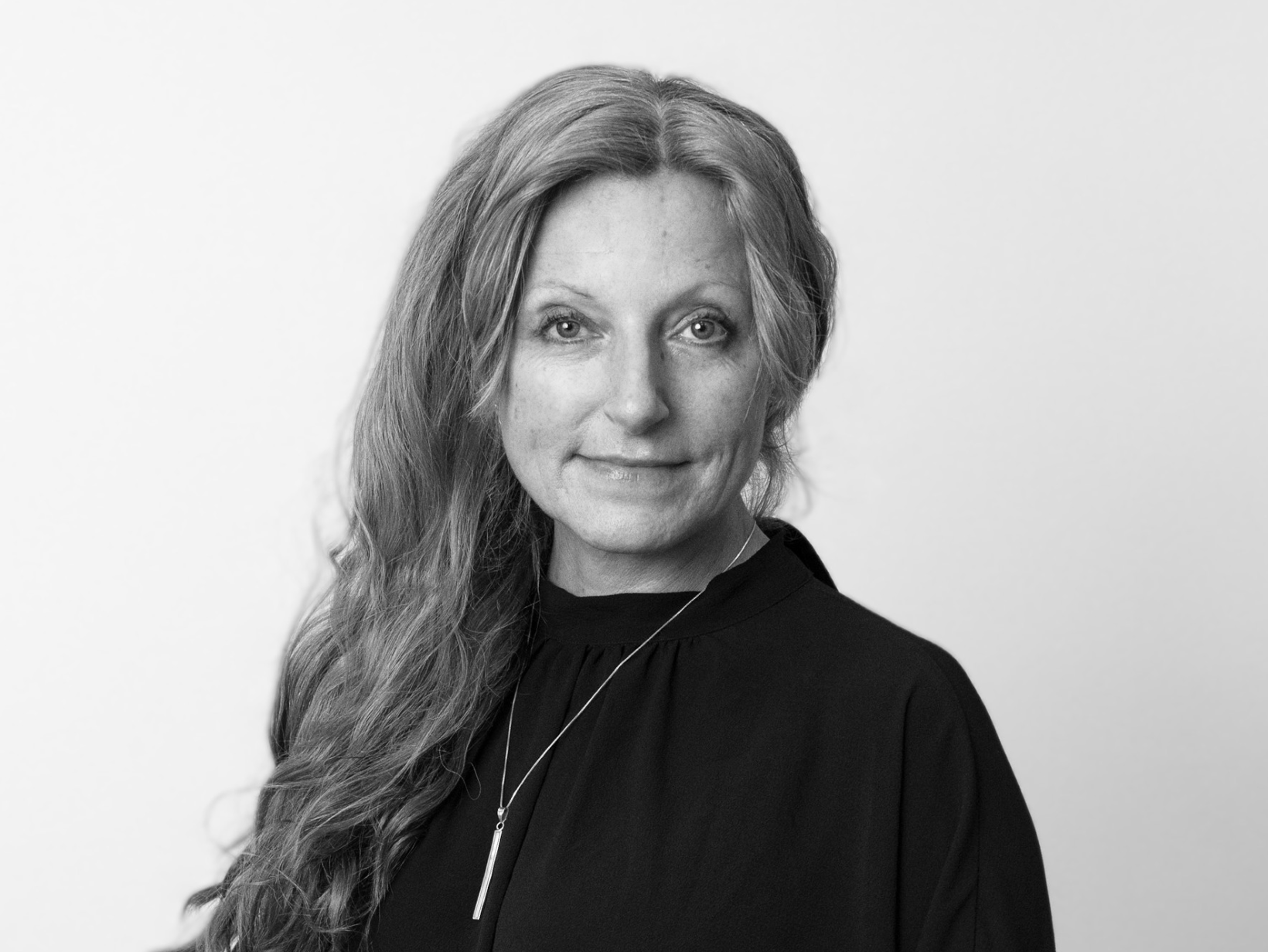 Black and white portrait of a woman with long wavy hair, wearing a dark top and a bar necklace.